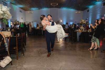 Groom carrying bride on the reception dance floor with cheering guests