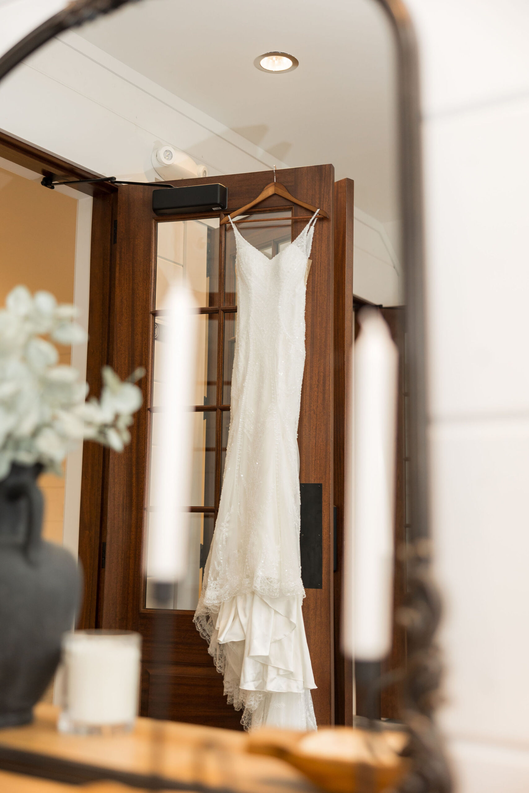 Beaded lace wedding gown hanging on dark wood doorway reflected in dressing mirror