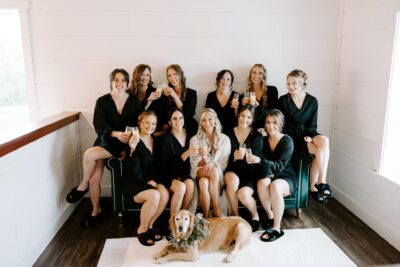 Bride and bridesmaids in black robes toasting champagne on the emerald sofa with pet dog