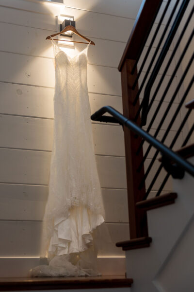 Wedding gown hanging under warm light fixture on shiplap wall beside staircase railing