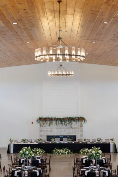 Grand reception head table set in front of stone fireplace with lush greenery garland and twin chandeliers
