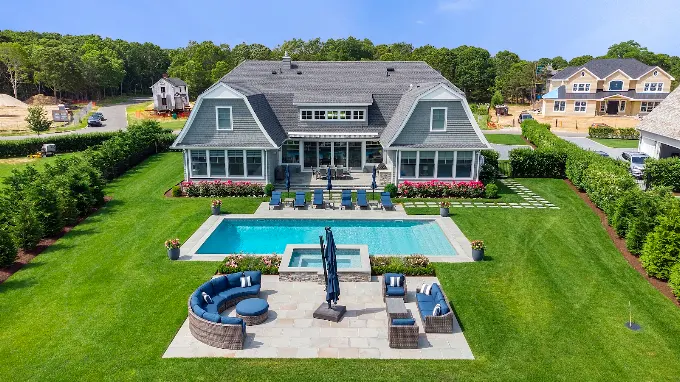 Aerial view of a grey cedar shingled home with a gambrel roof, pool, spa and bluestone patio