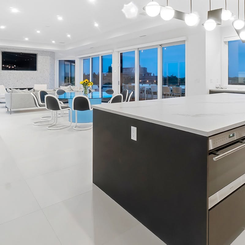 Open plan kitchen with a waterfall quartz island, wall ovens and large format porcelain flooring, opening onto a dining area and sliding glass doors