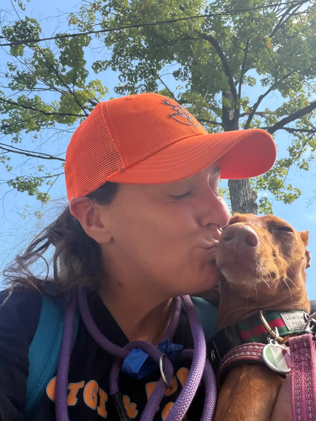 Affectionate kiss on the nose with brown rescue pup in orange hat under trees — Super Gigi pet sitting and dog walking in Bergen County, NJ