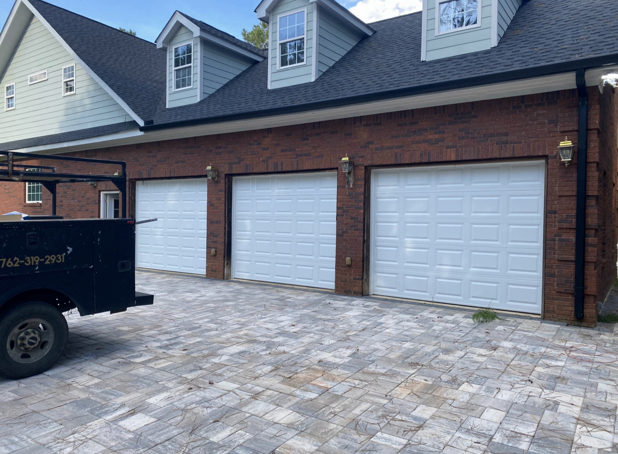 Three white raised-panel garage doors installed on a brick residence with paved driveway by All Access Garage Doors