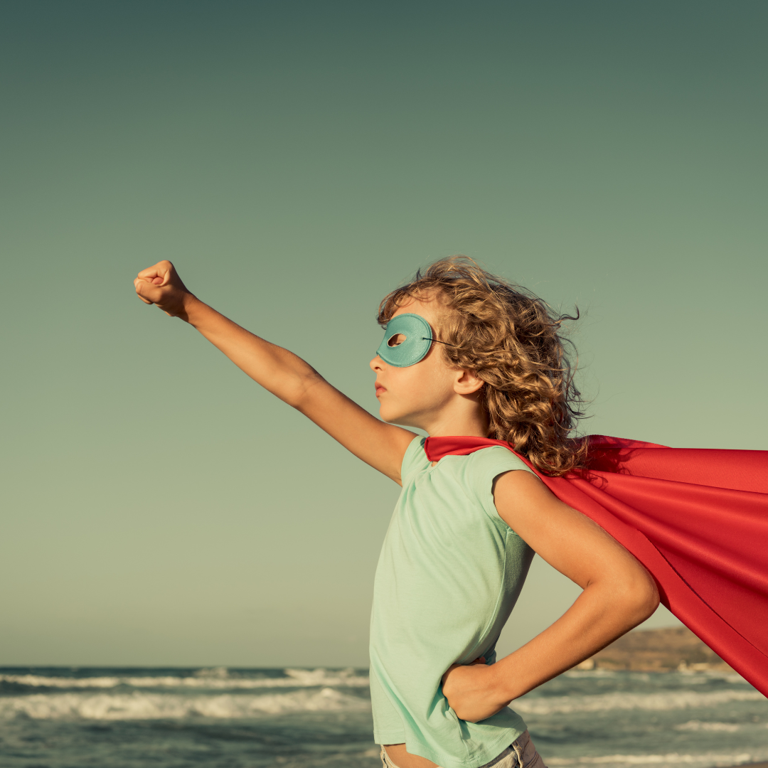 Child wearing superhero mask and cape standing confidently on the beach