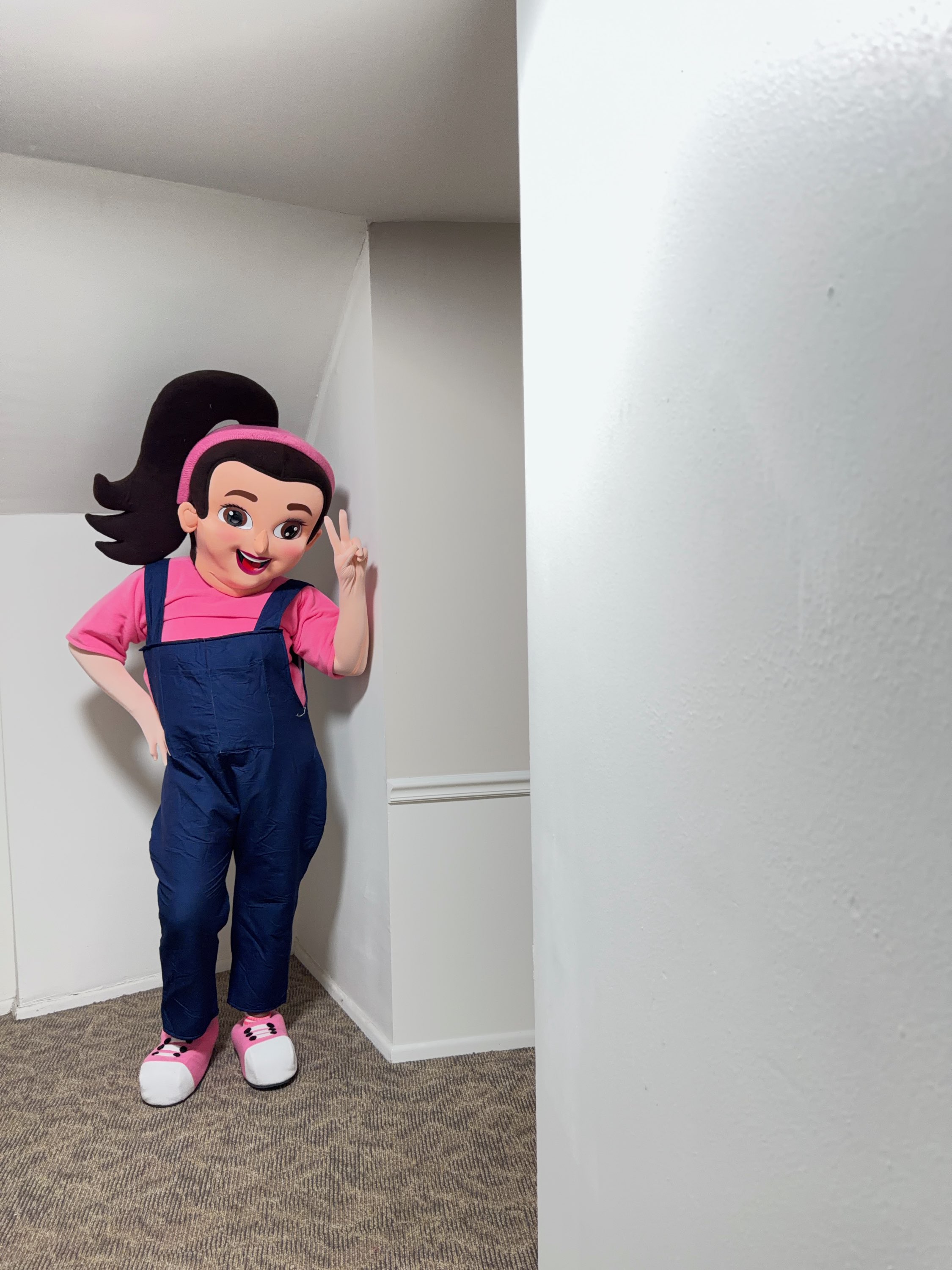 Kaleahs mascot character wearing navy overalls and pink top flashing peace sign