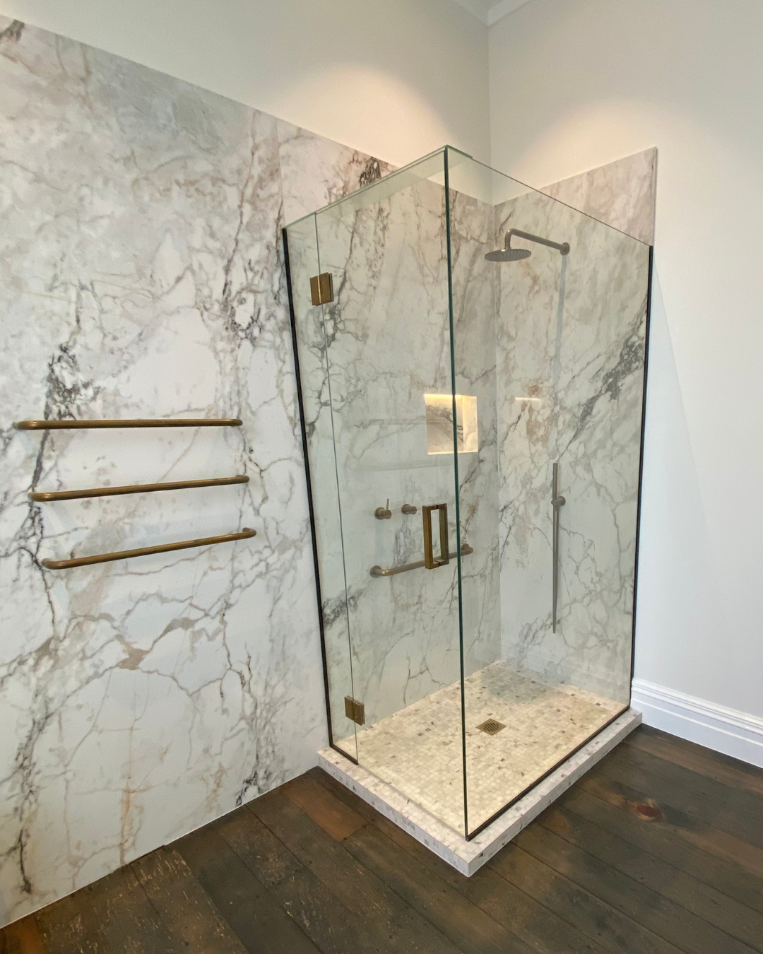 Marble feature wall and glass shower enclosure with brass hardware and wood flooring.