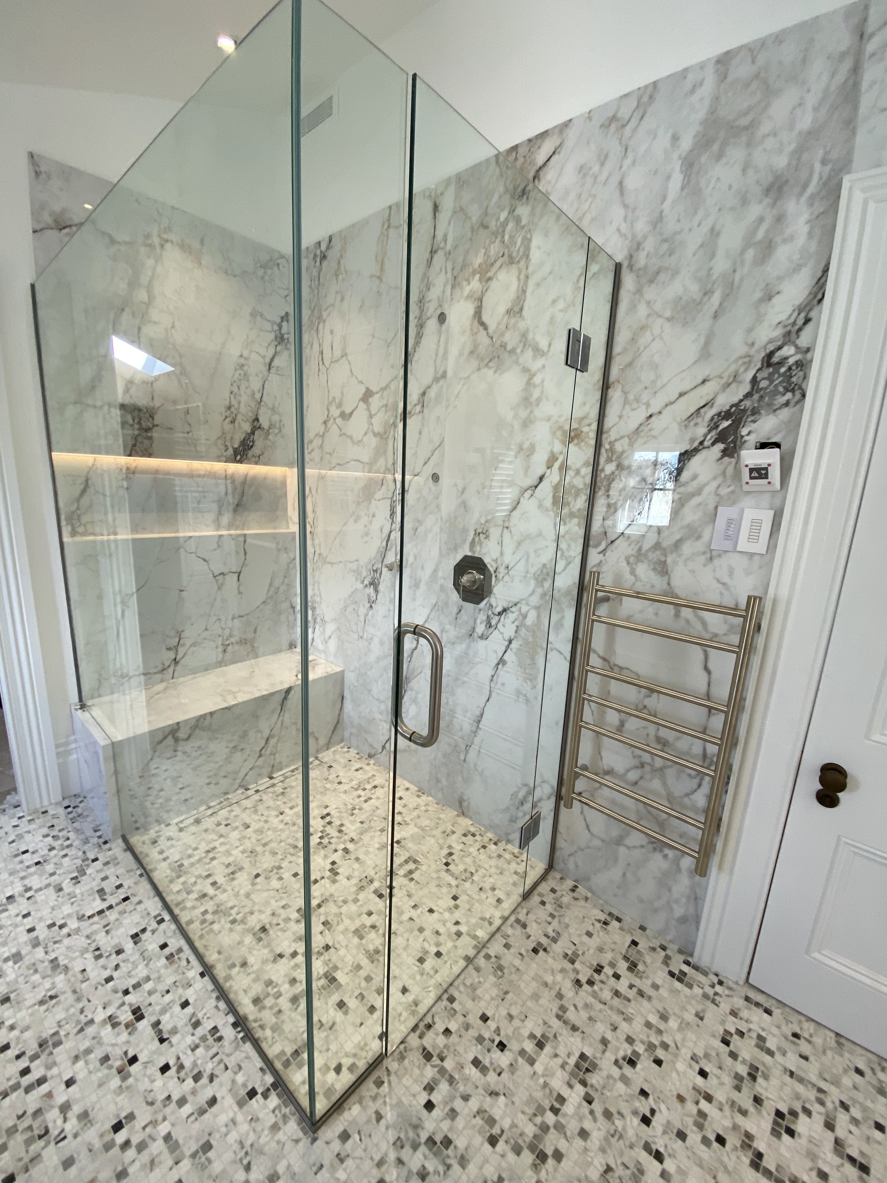 Custom glass walk-in shower with marble slab walls, illuminated niche, integrated marble bench, and mosaic floor.