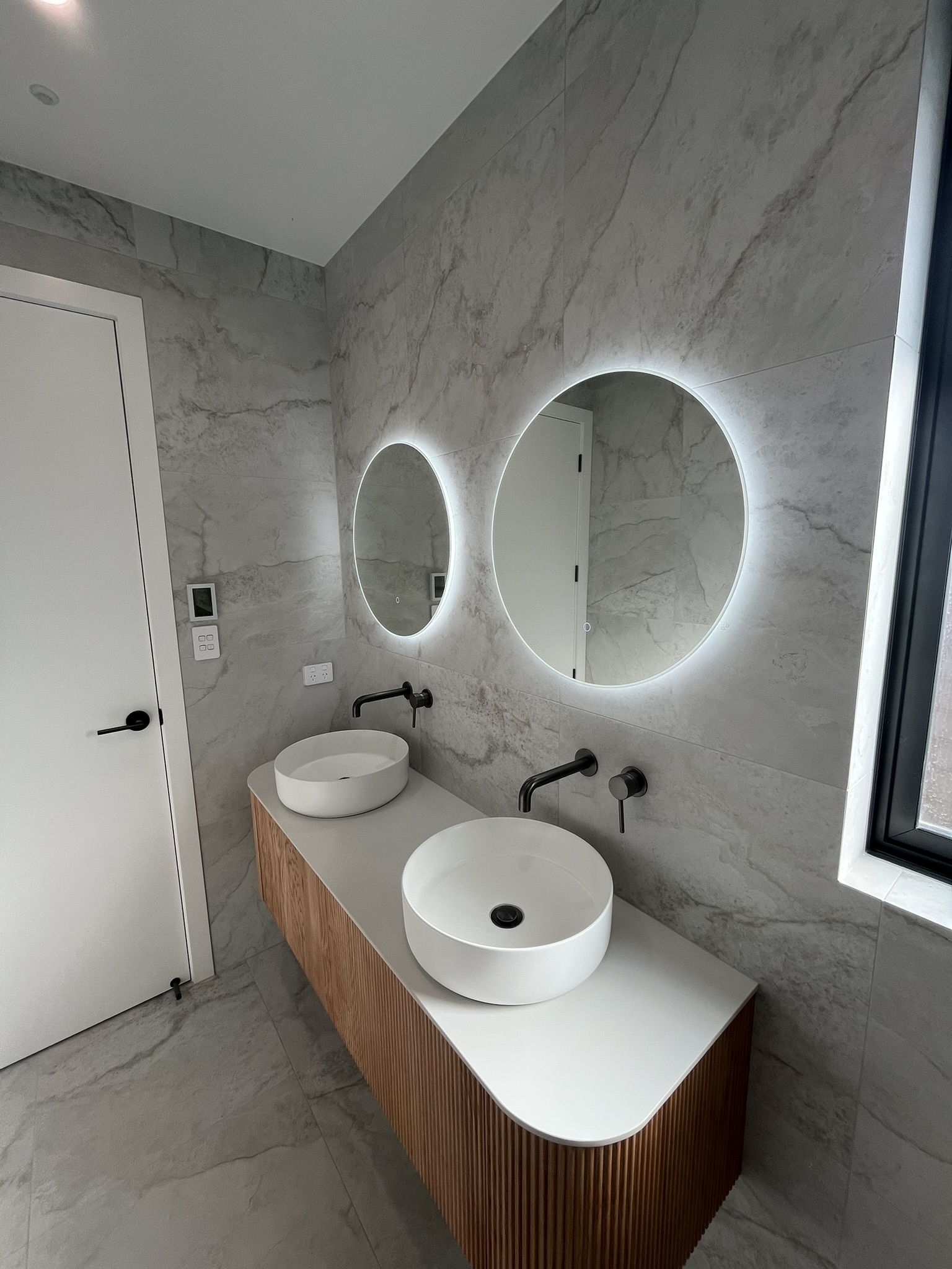 Luxury bathroom vanity feature wall with grey stone tiles, fluted timber vanity, and dual backlit circular mirrors.
