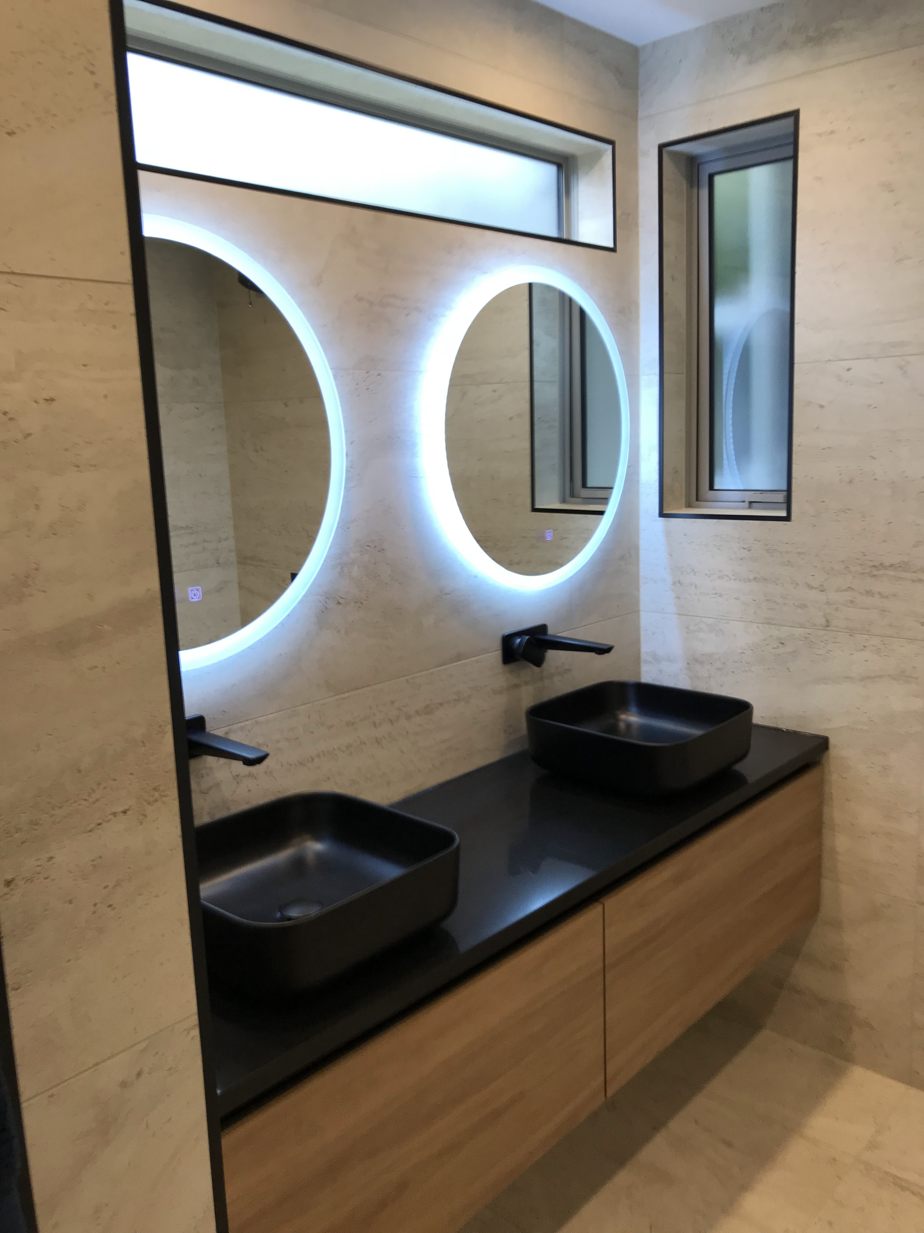 Travertine style wall tiling behind floating timber vanity and dual backlit circular mirrors.
