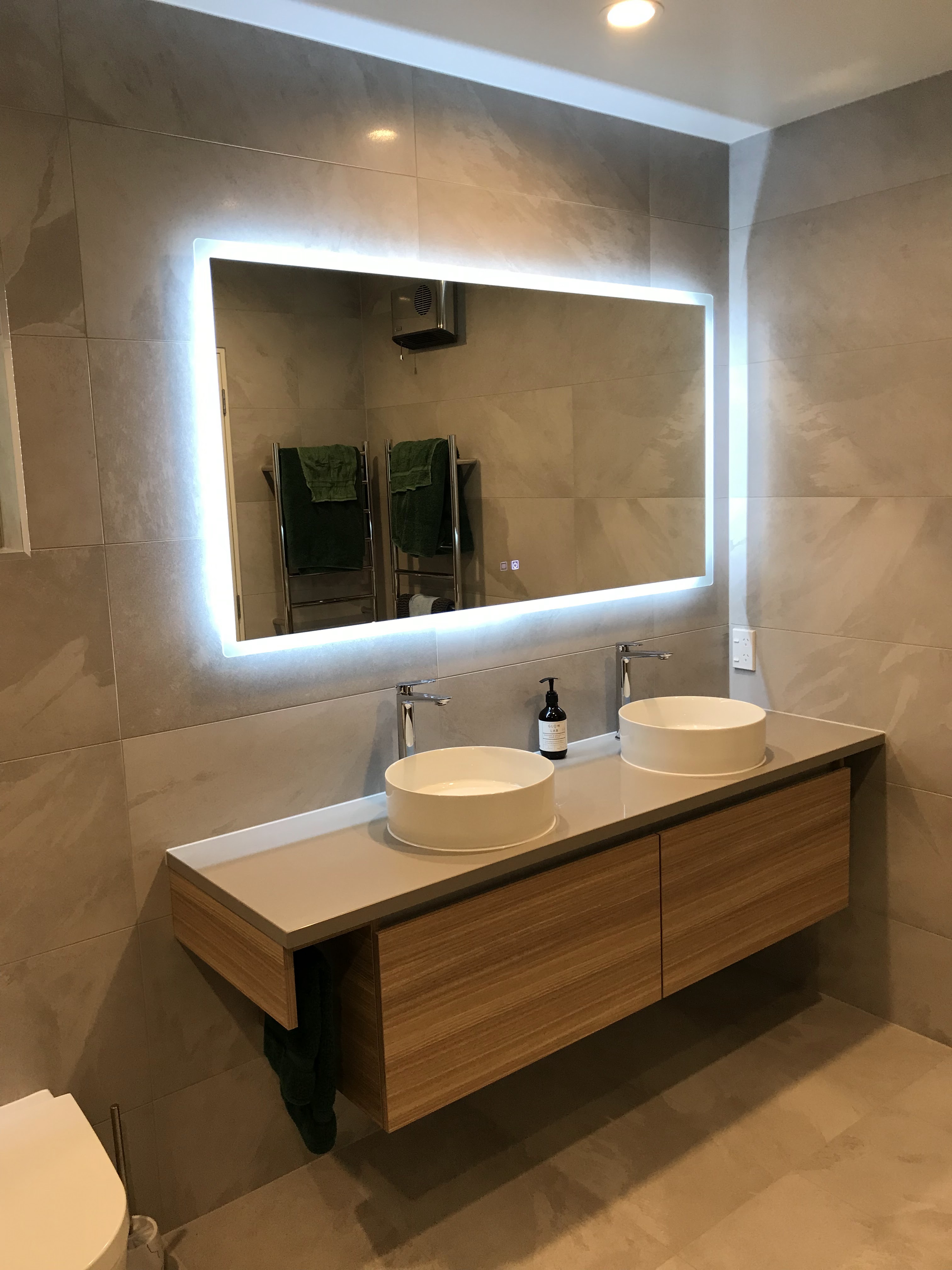 Contemporary bathroom wall tiling with floating timber double vanity, dual vessel basins, and illuminated mirror.