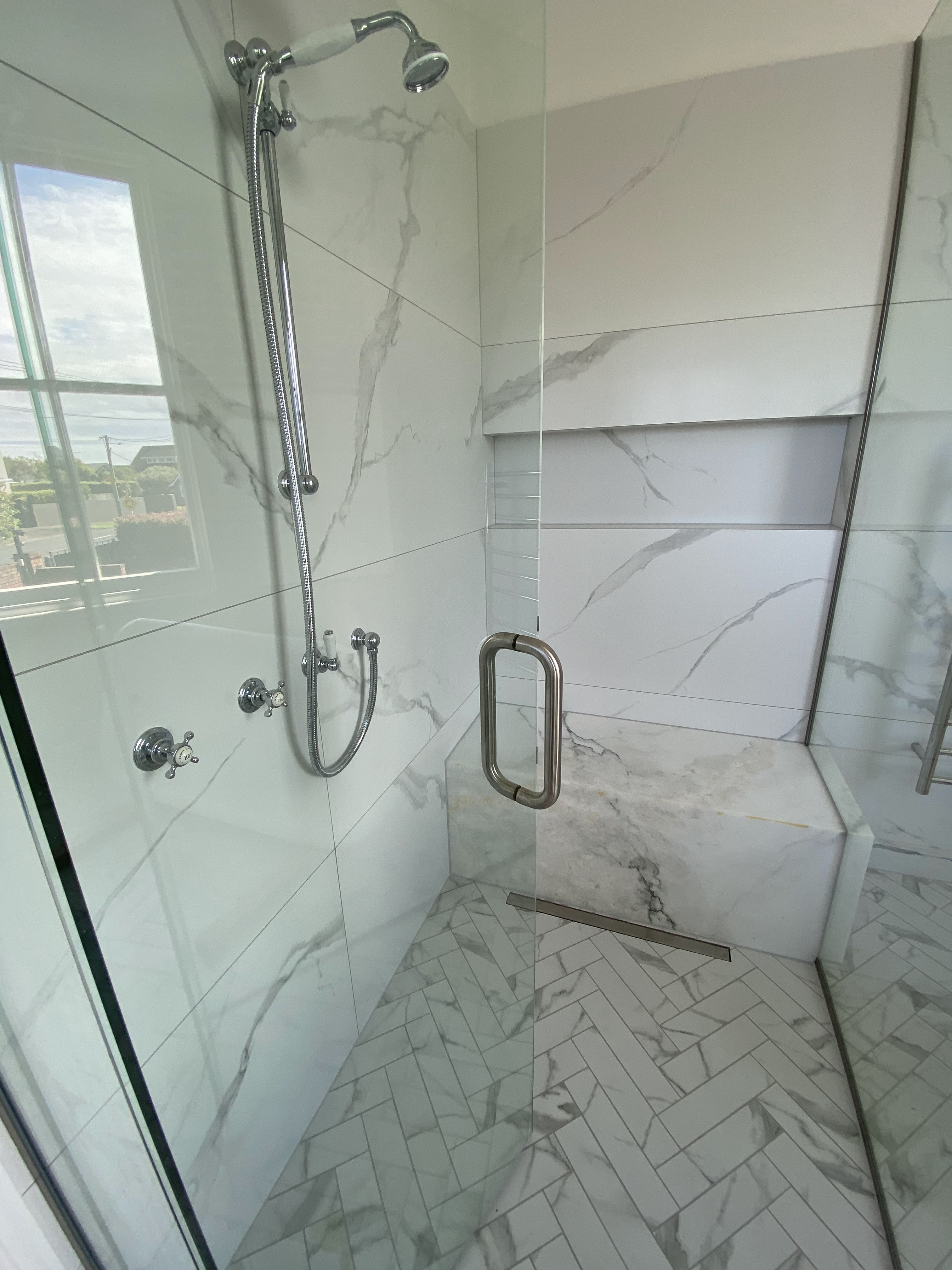 Custom marble walk-in shower with integrated marble bench, herringbone tiled floor, and recessed shelf.