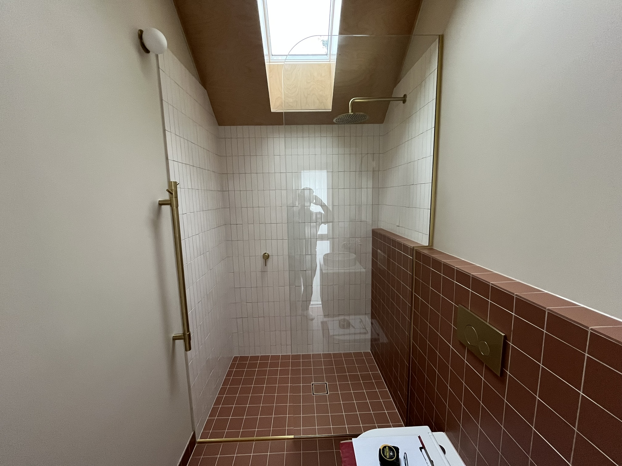 Architectural walk-in shower with terracotta tiles, vertical white subway walls, brass hardware, and skylight.