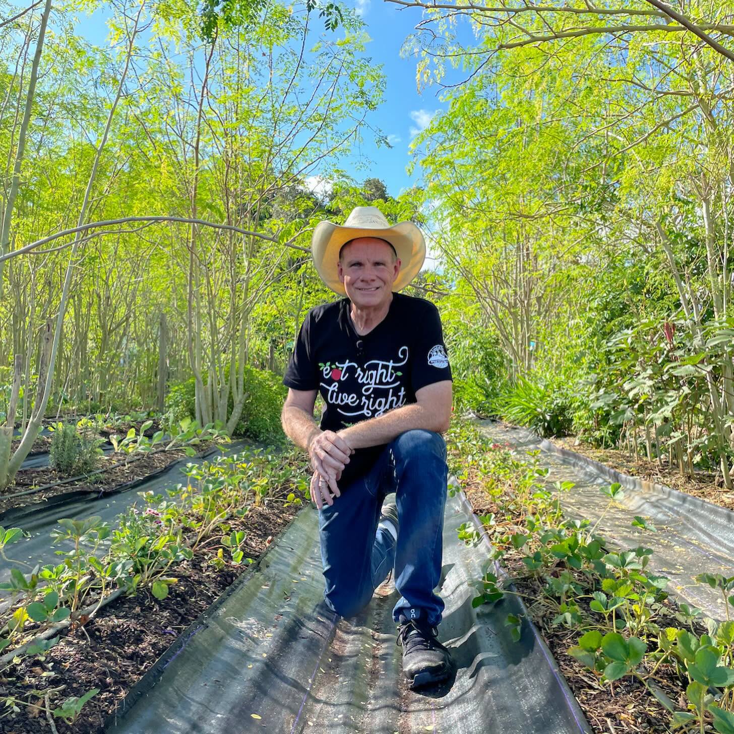 Farm founder kneeling in organic moringa and strawberry crop rows wearing 'Eat Right Live Right' shirt and cowboy hat
