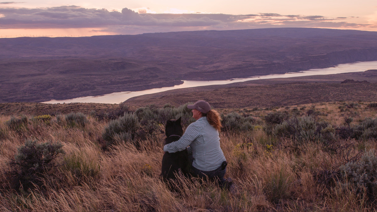 Trainer embracing black dog overlooking river valley sunset