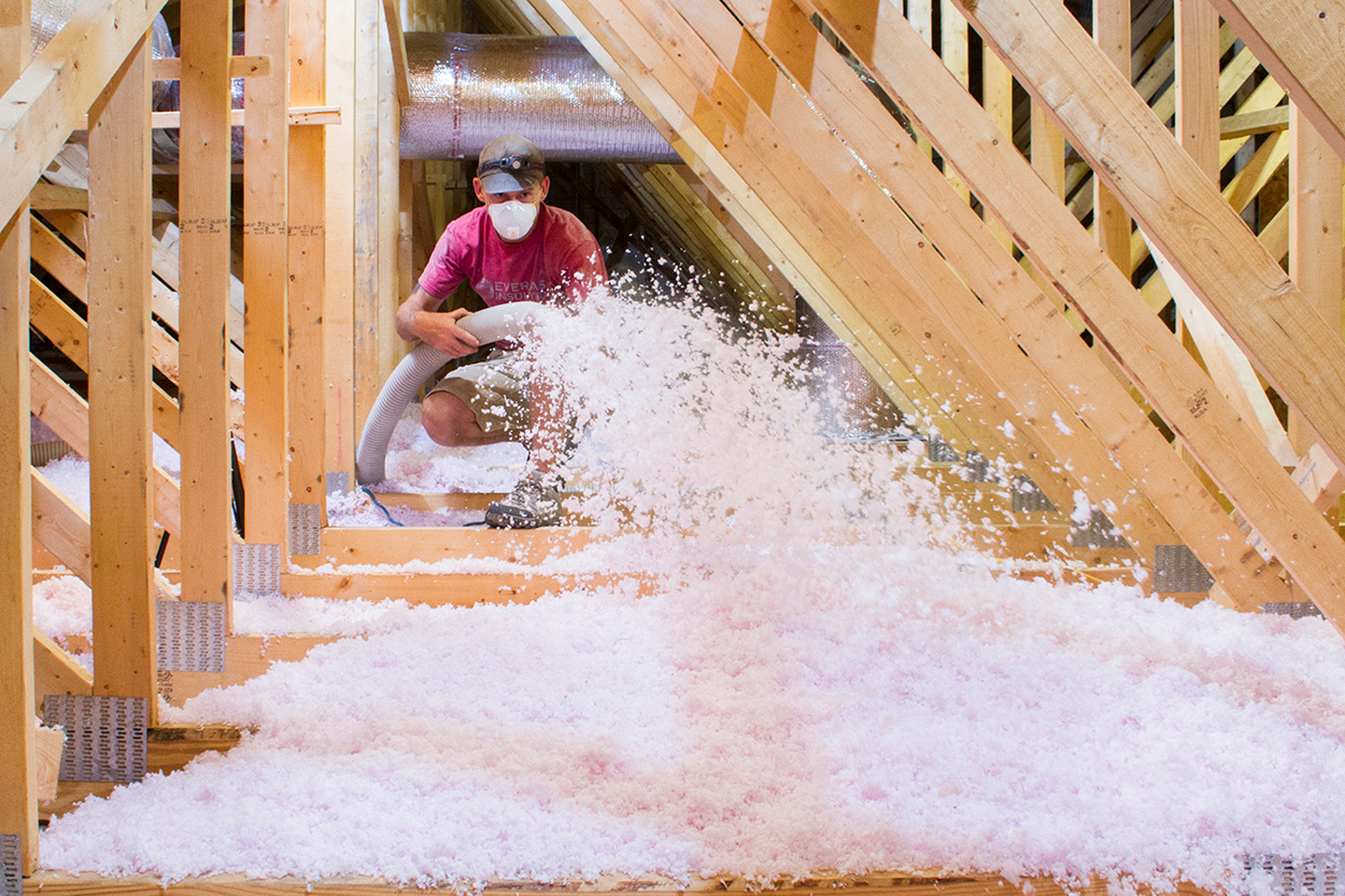 Technician in protective gear blowing pink loose-fill insulation across roof trusses in new construction attic