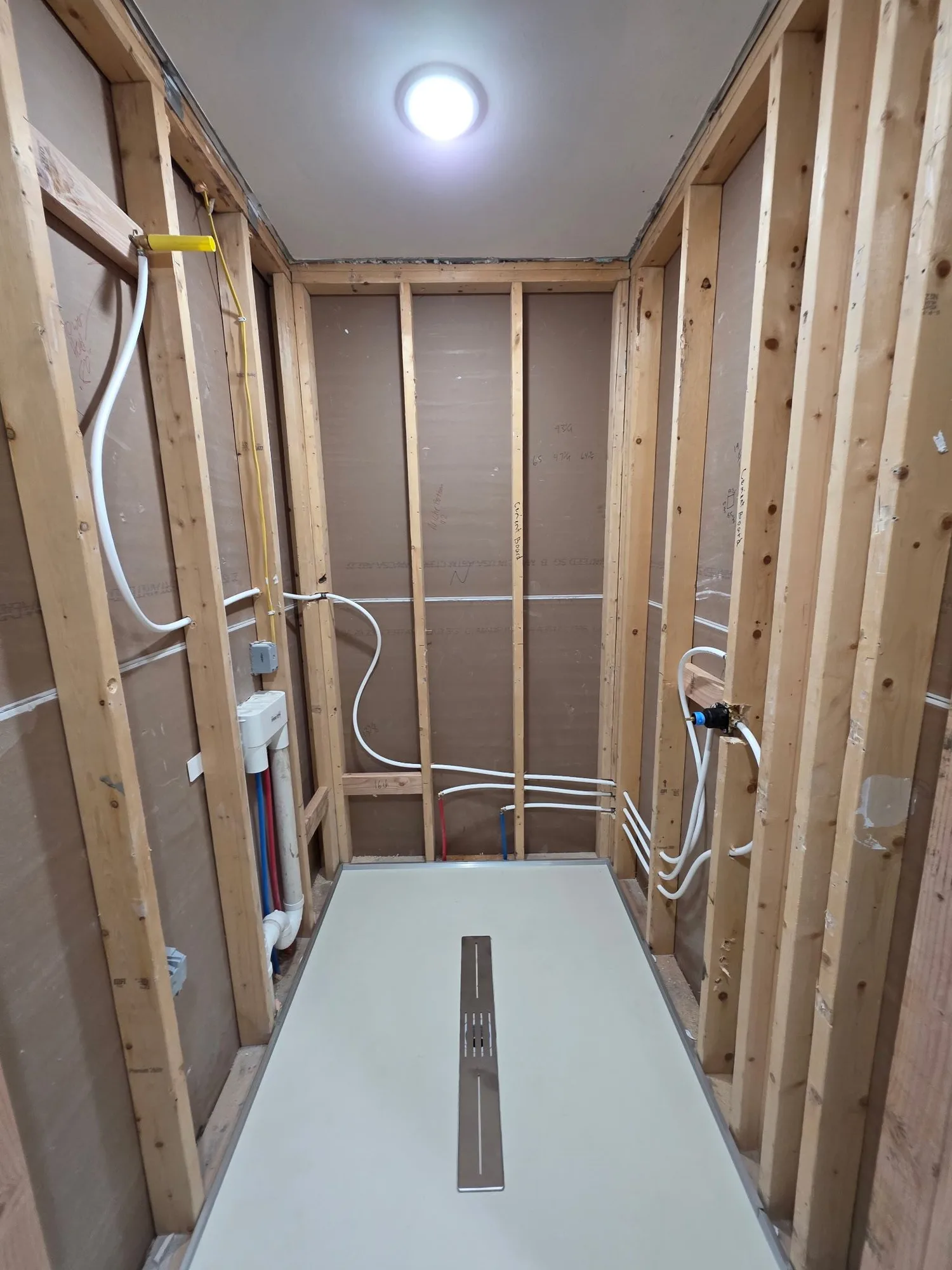 H&H Home & Bath Renovations in-progress shower remodel with exposed wood studs, rough-in plumbing, PEX supply lines, and newly installed shower pan with linear drain