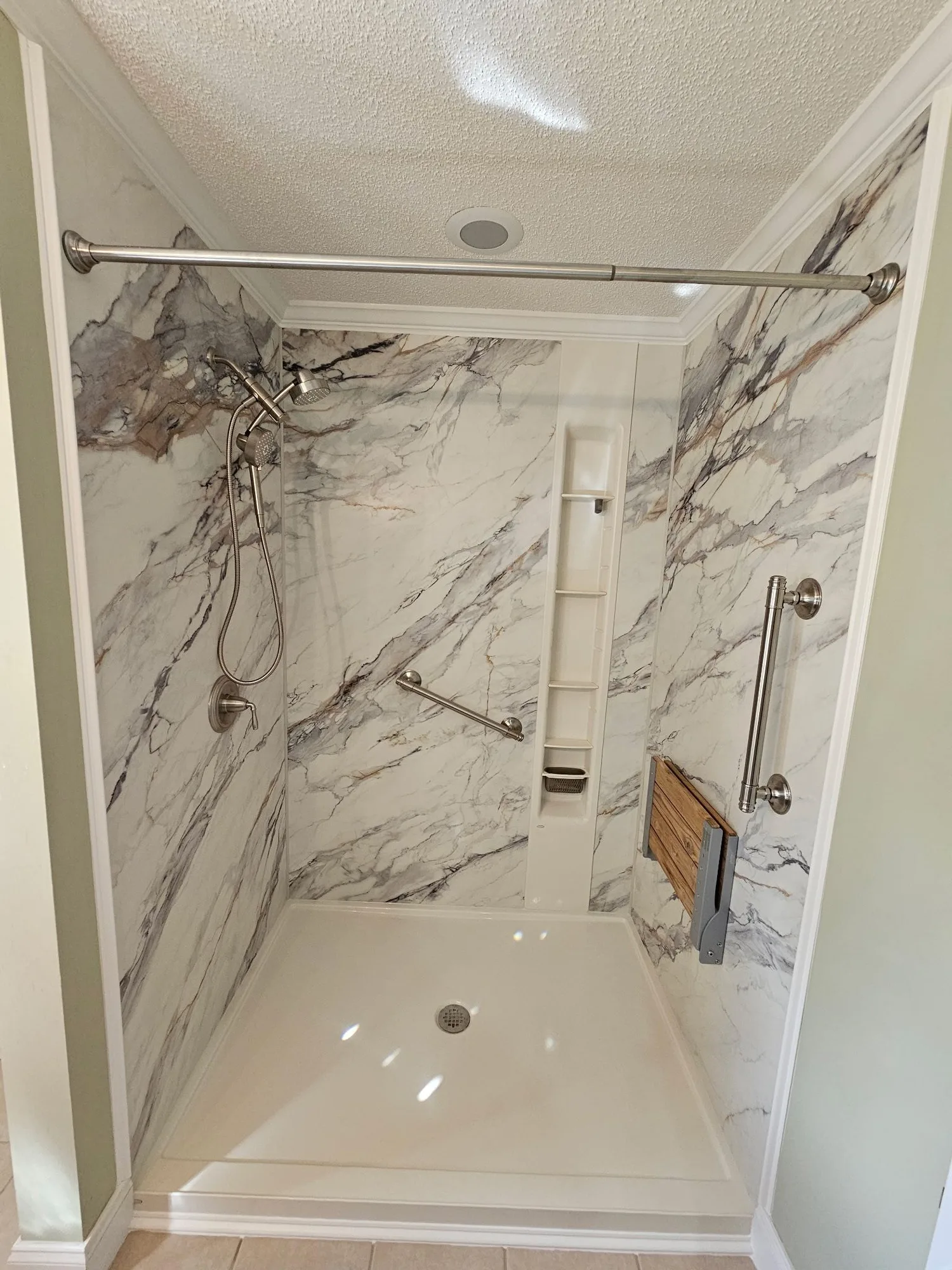 H&H Home & Bath Renovations completed custom walk-in shower remodel featuring marble-pattern wall panels, handheld shower wand, safety grab bars, fold-down bench seat, and built-in shelving niche