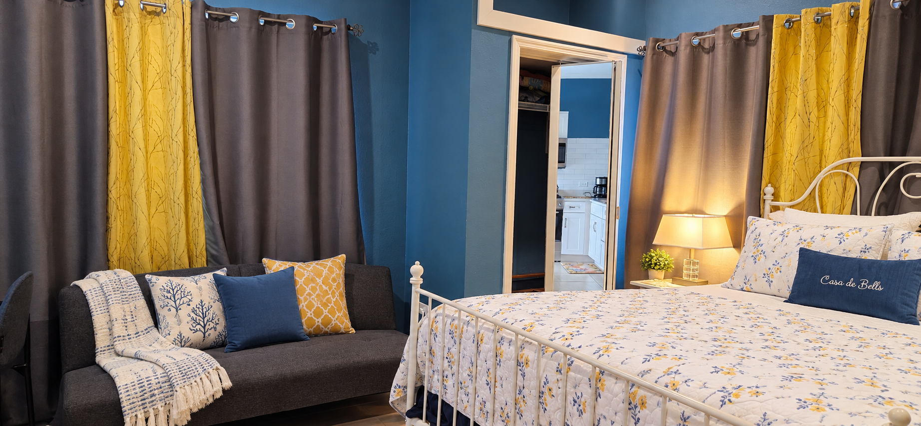 Studio suite with white wrought-iron bed, sofa seating area, and entrance to kitchen