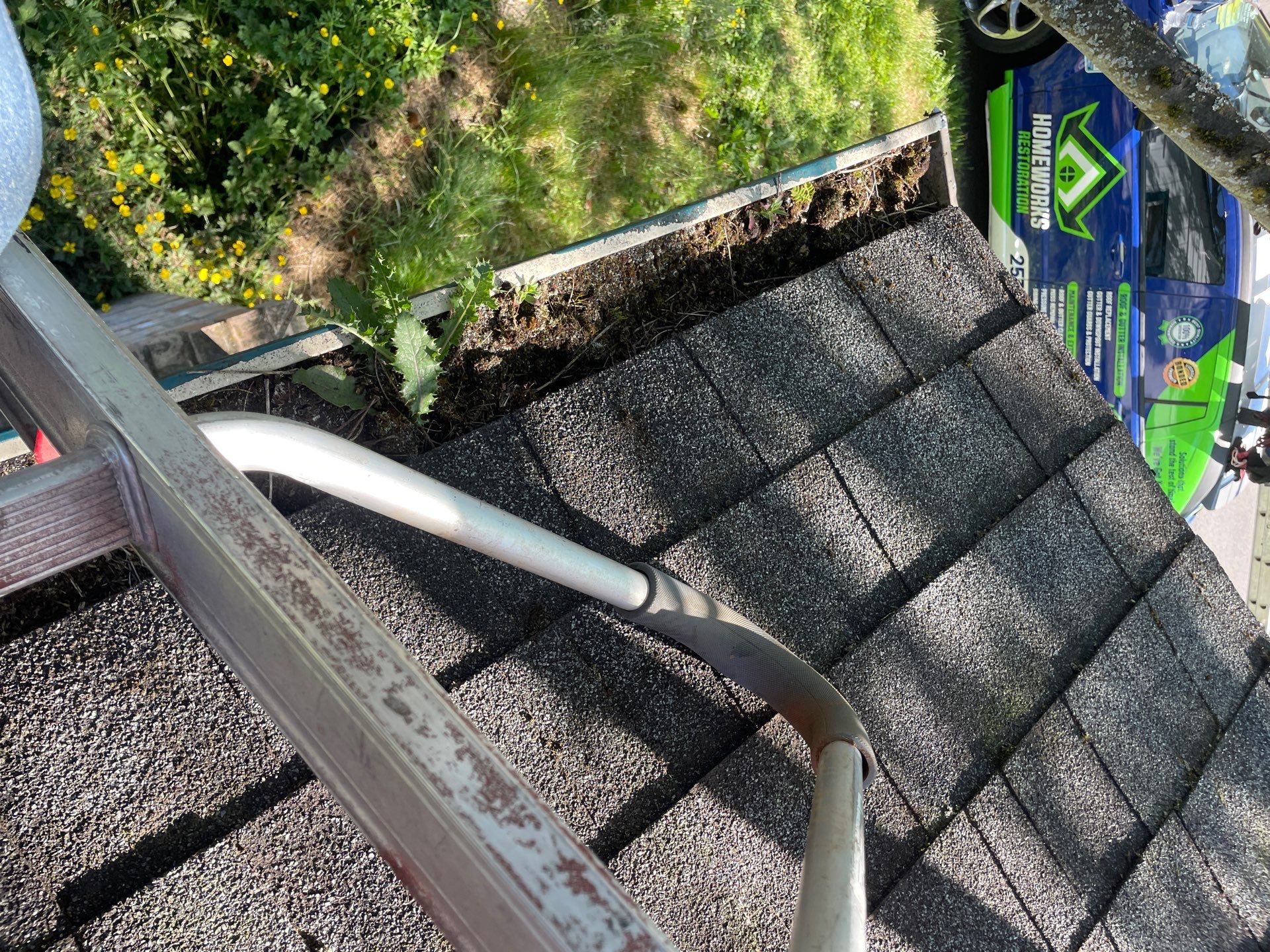 Before gutter cleaning — gutter channel severely clogged with packed mud, compost, and weeds sprouting along roofline with Homeworks Restoration service vehicle below