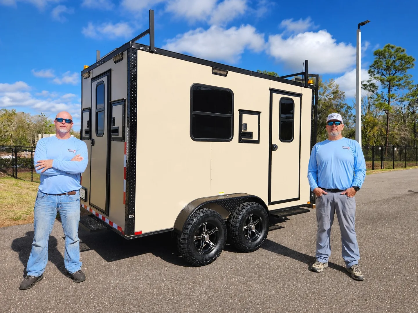 Two team members in matching blue shirts standing beside a tan enclosed utility trailer with a roof rack and off-road tires.