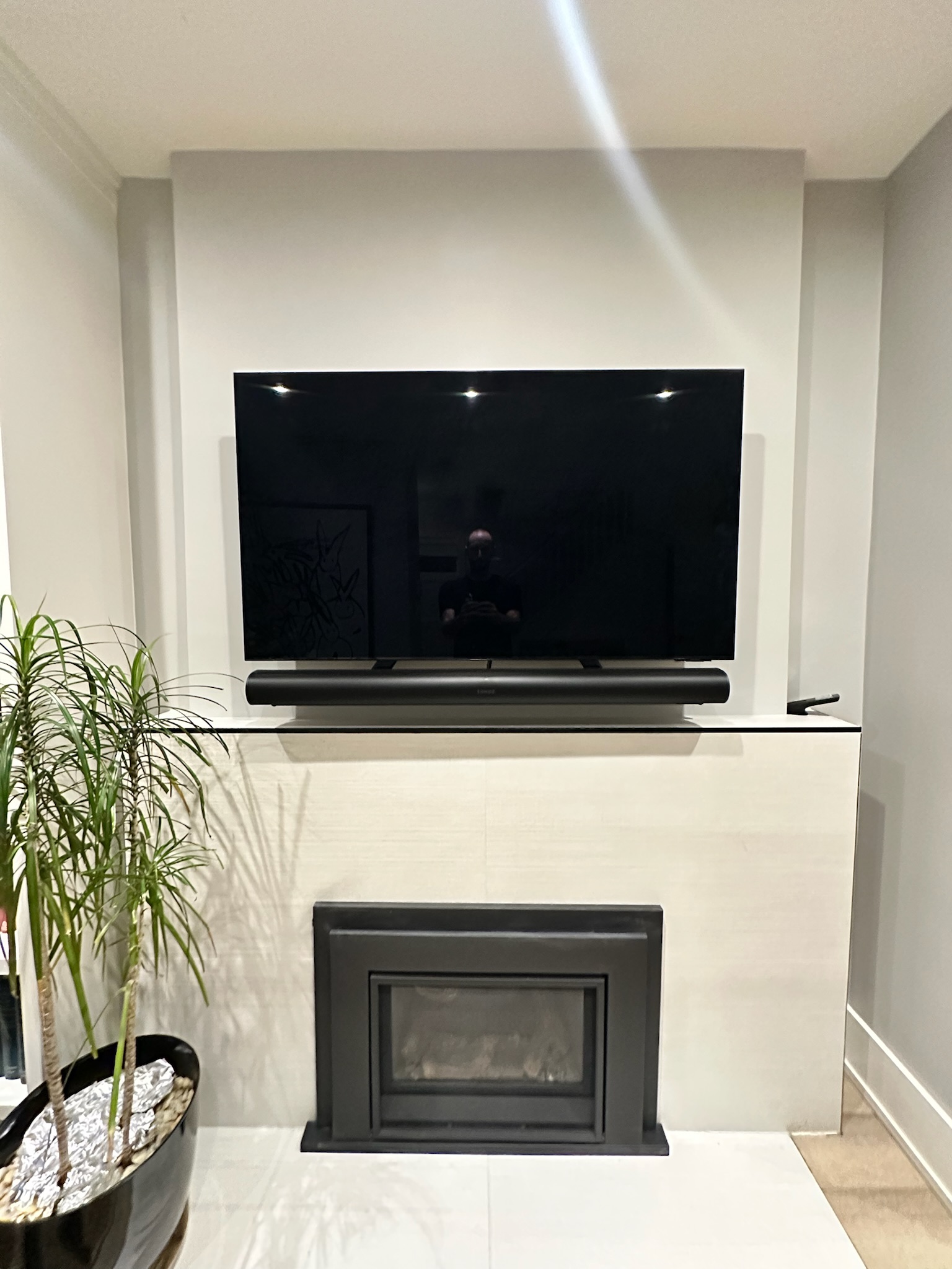 Mount Masters OLED TV with floating soundbar mounted flush above modern white tiled fireplace and black inset mantel