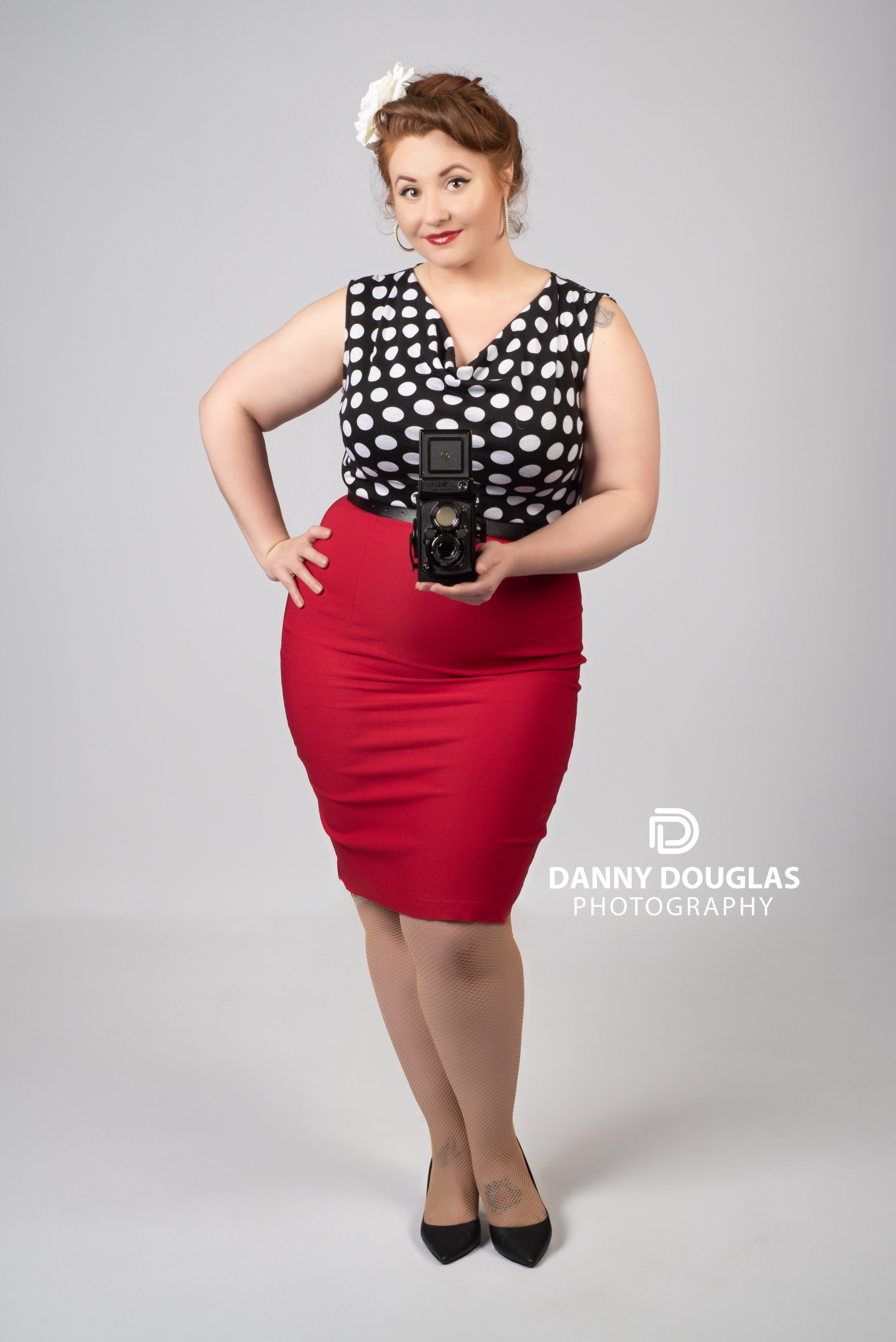Pin-up portrait in a black and white polka dot top and red pencil skirt holding a vintage twin-lens camera