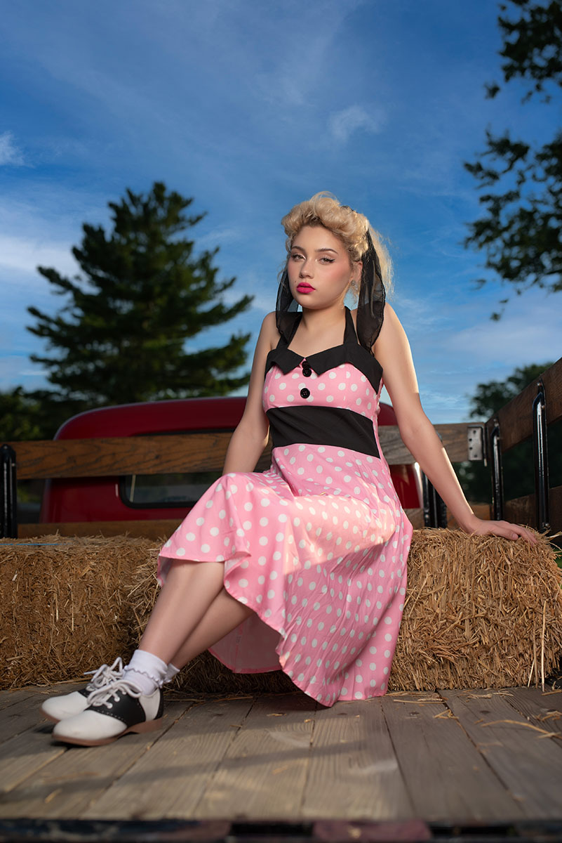 Pin-up model in a pink polka dot dress and saddle shoes on vintage truck hay bales