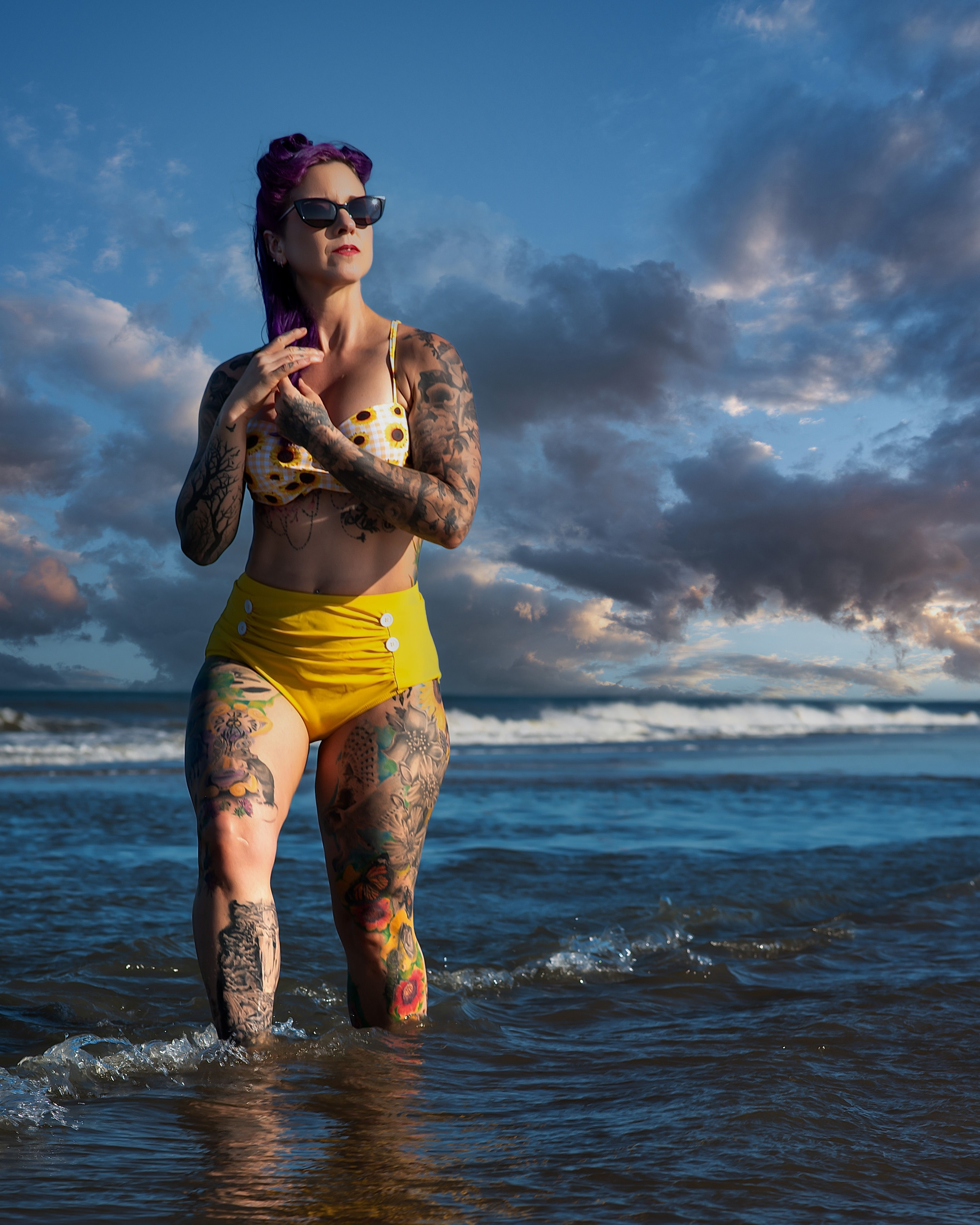 Tattooed pin-up model wading in the ocean surf in a high-waisted yellow bikini and sunglasses