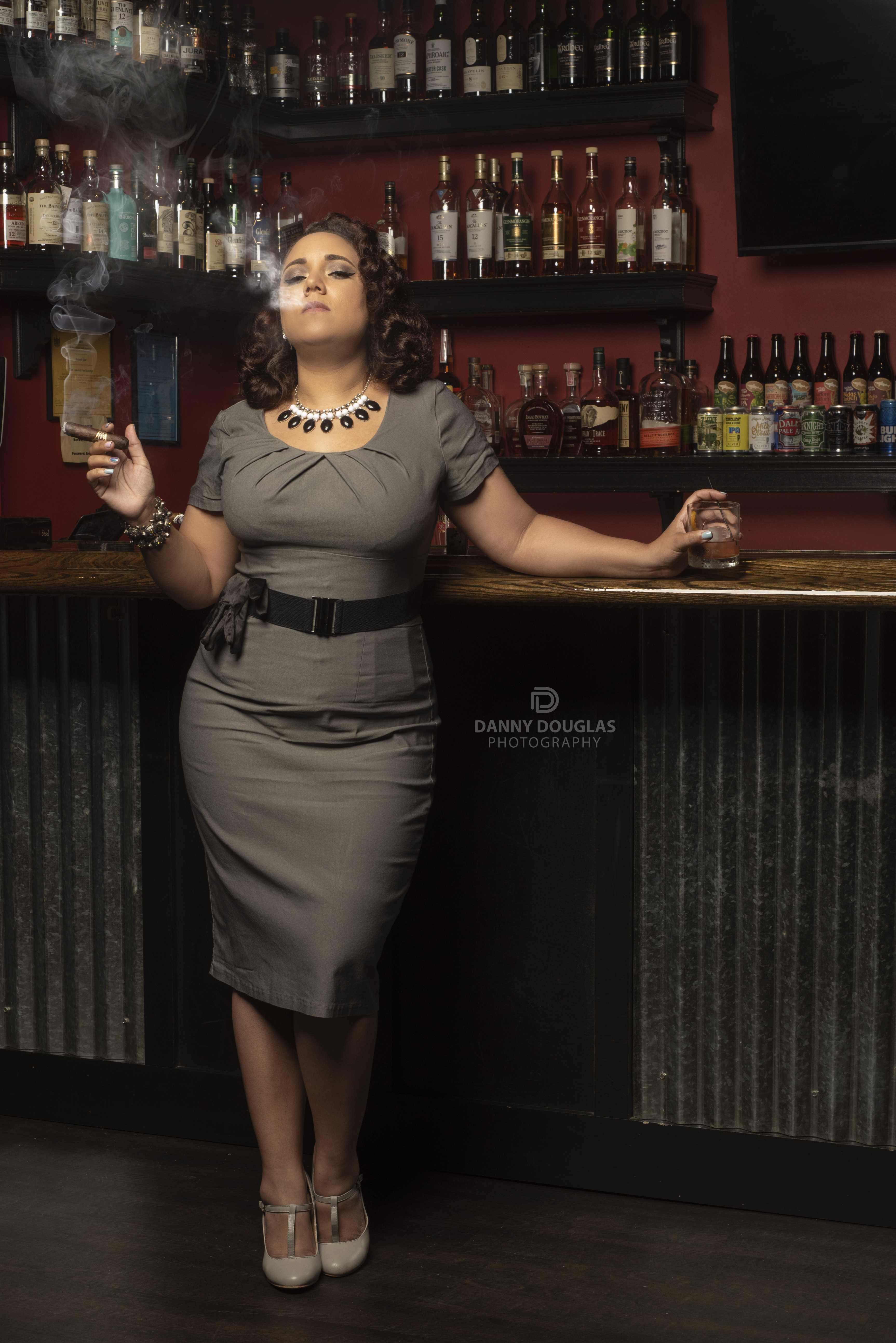 Vintage glam portrait of a woman in a grey pencil dress at a whiskey bar with cigar smoke
