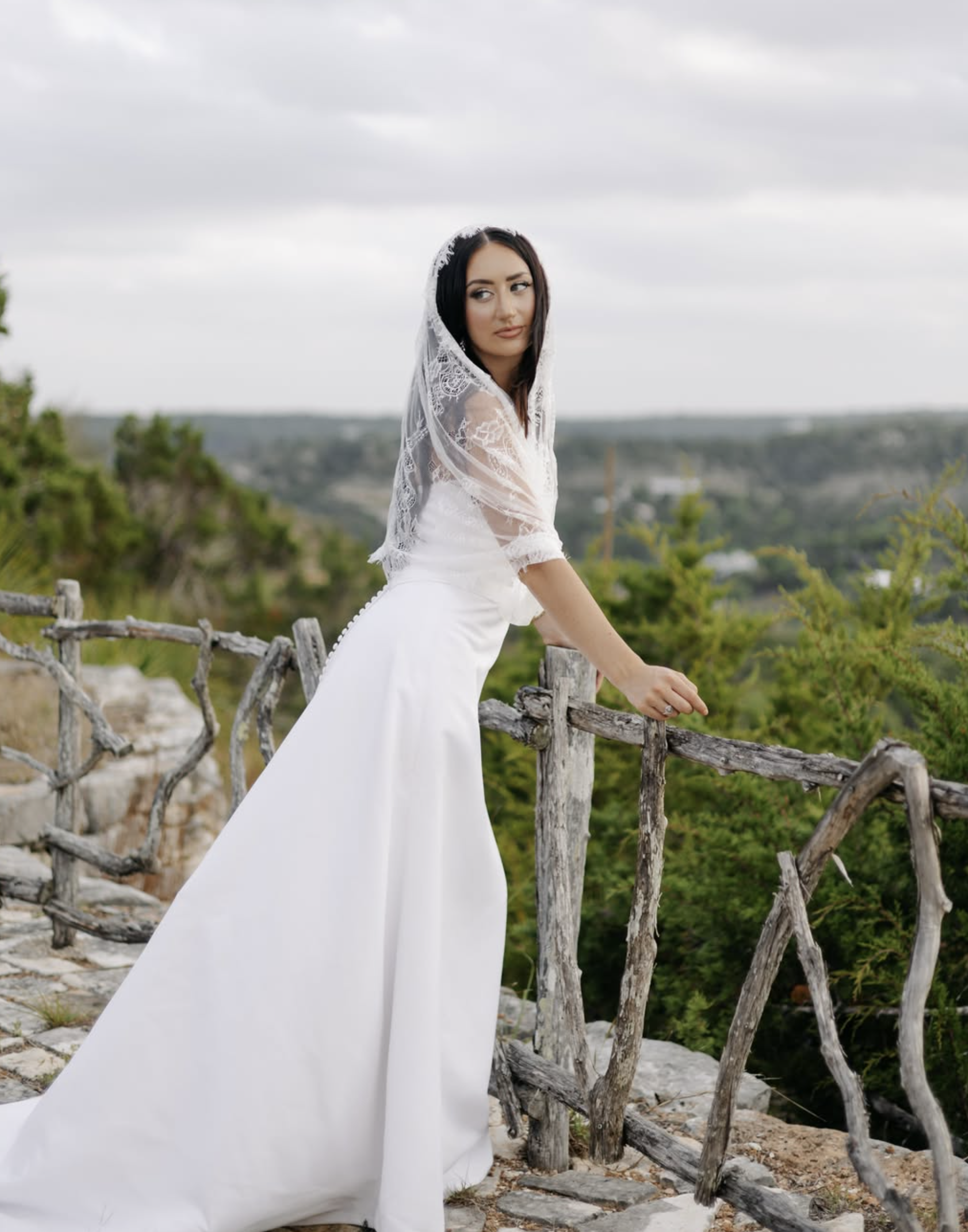 Bride wearing lace veil leaning gently against the cedar overlook railing with Hill Country vistas