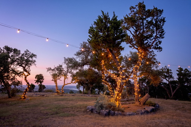Outdoor hillside grounds under bistro string lights wrapped around Texas oak and cedar trees at twilight overlooking Canyon Lake