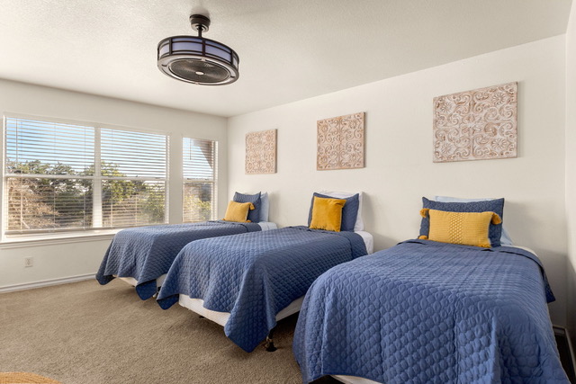 Triple-bed guest room with cozy blue quilts, accent pillows, ceiling fan, and hillside window light for group retreat lodging