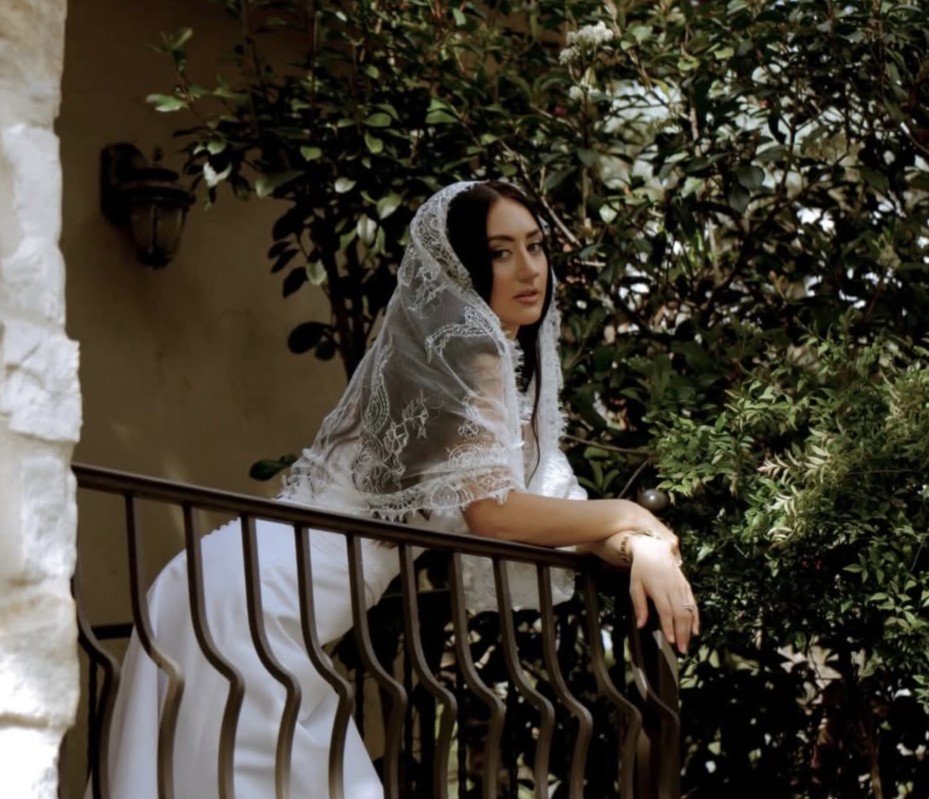 Bride resting gracefully against stone and iron balcony railing draped in lace veil surrounded by lush greenery