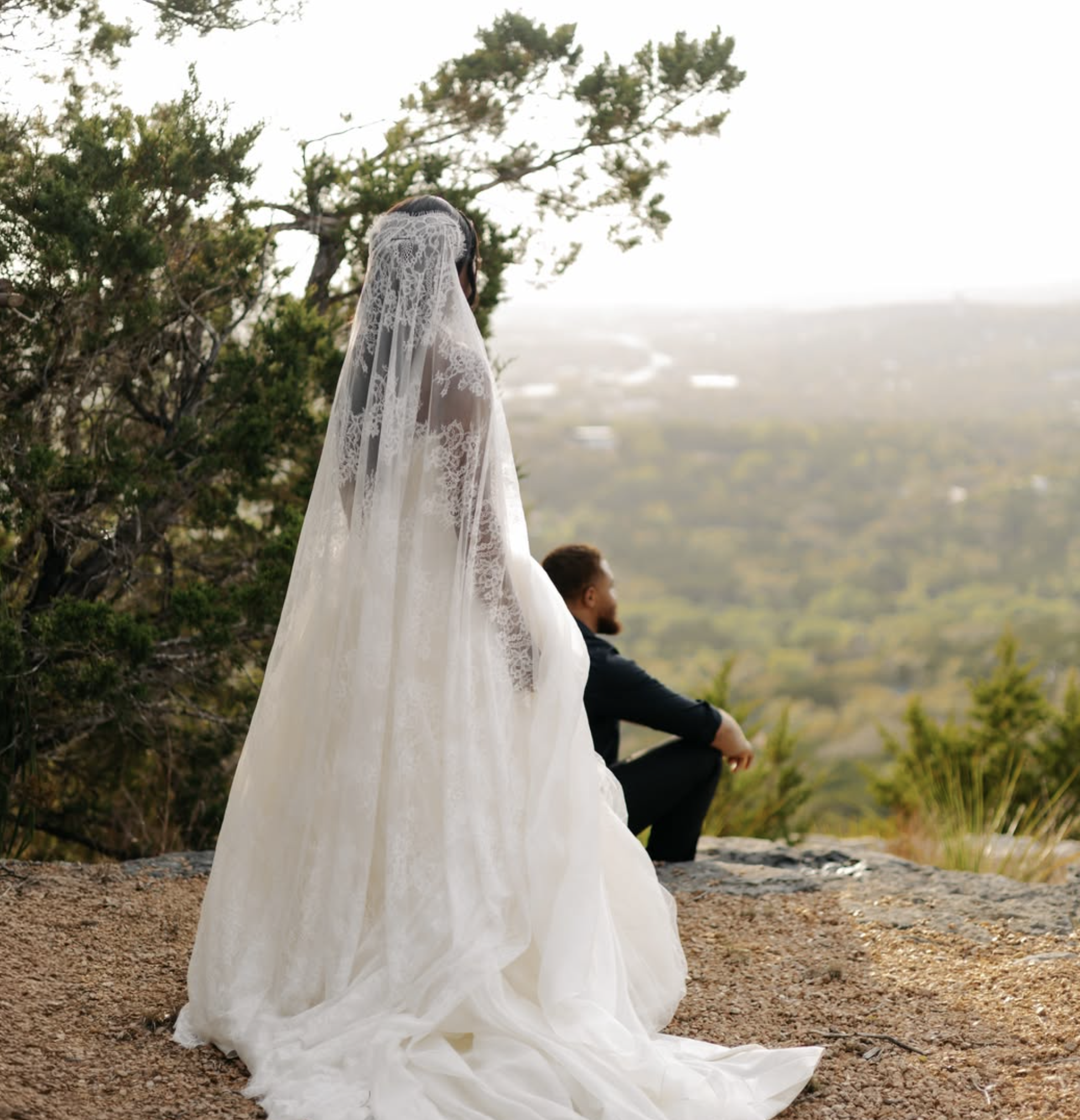 Bride in cathedral lace veil standing at the rocky overlook with groom seated looking over Canyon Lake hills