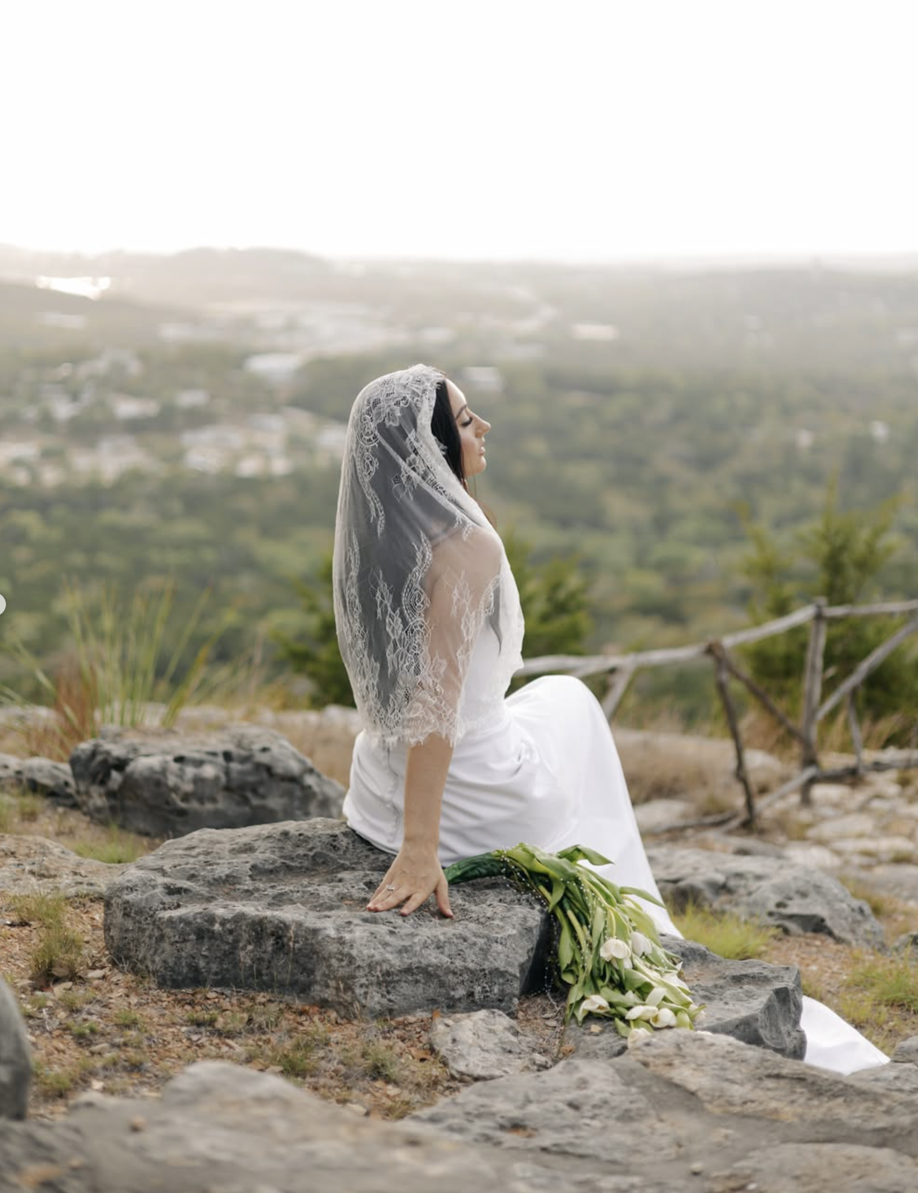 Bride in lace mantilla veil seated serenely on limestone rocks with white bridal bouquet above Hill Country vistas
