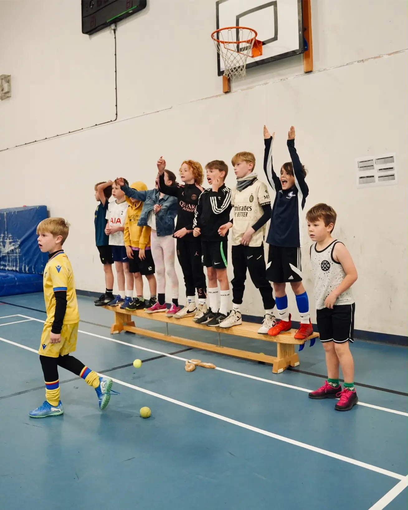 Children lined up in sports kit cheering with hands raised during an indoor holiday camp session in Esher
