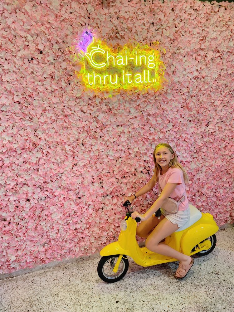 Girl smiling on yellow scooter at Chai-ing thru it all flower wall