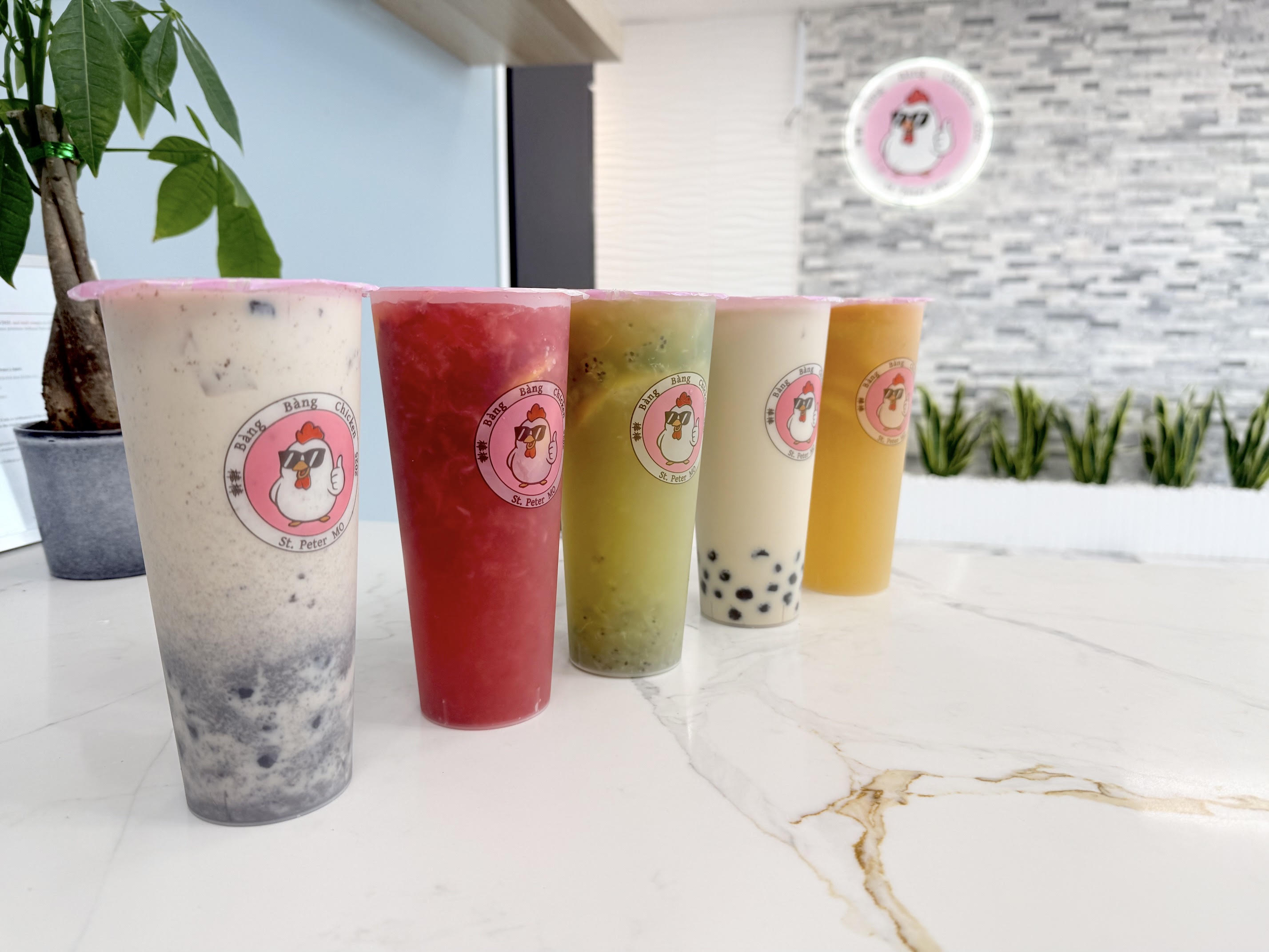 Five colorful boba and fruit drinks on marble counter in front of pink neon chicken logo