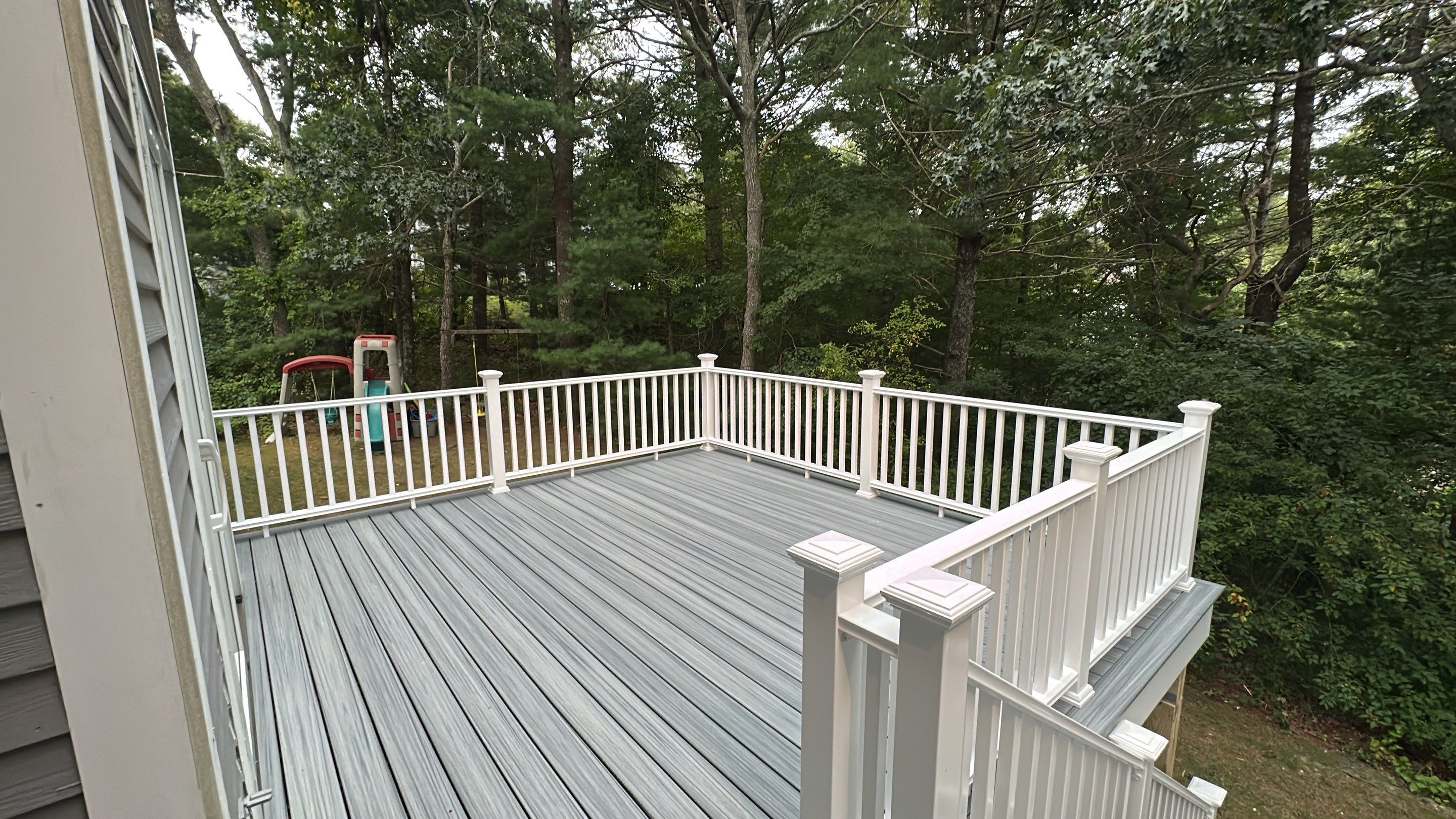 High angle view across newly completed grey composite deck surface surrounded by white vinyl railings overlooking private woods