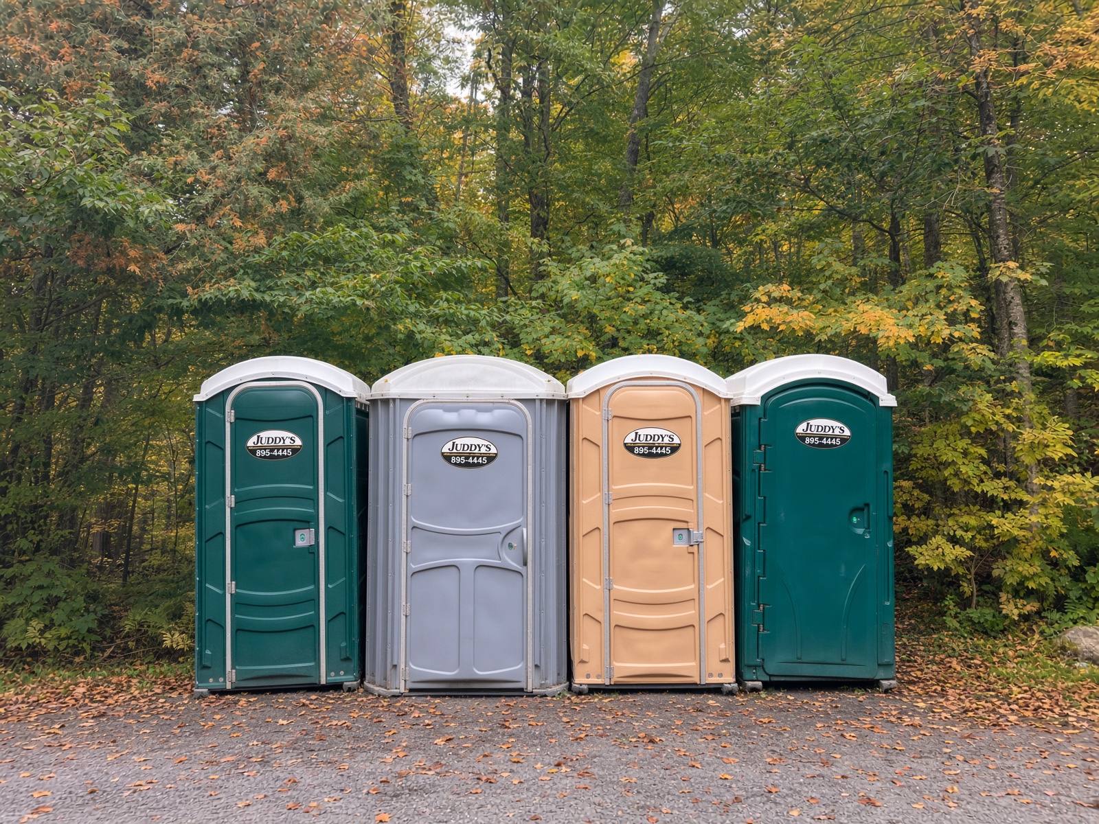 Four clean Juddy's portable toilets in green, grey, and tan lined up against Vermont trees