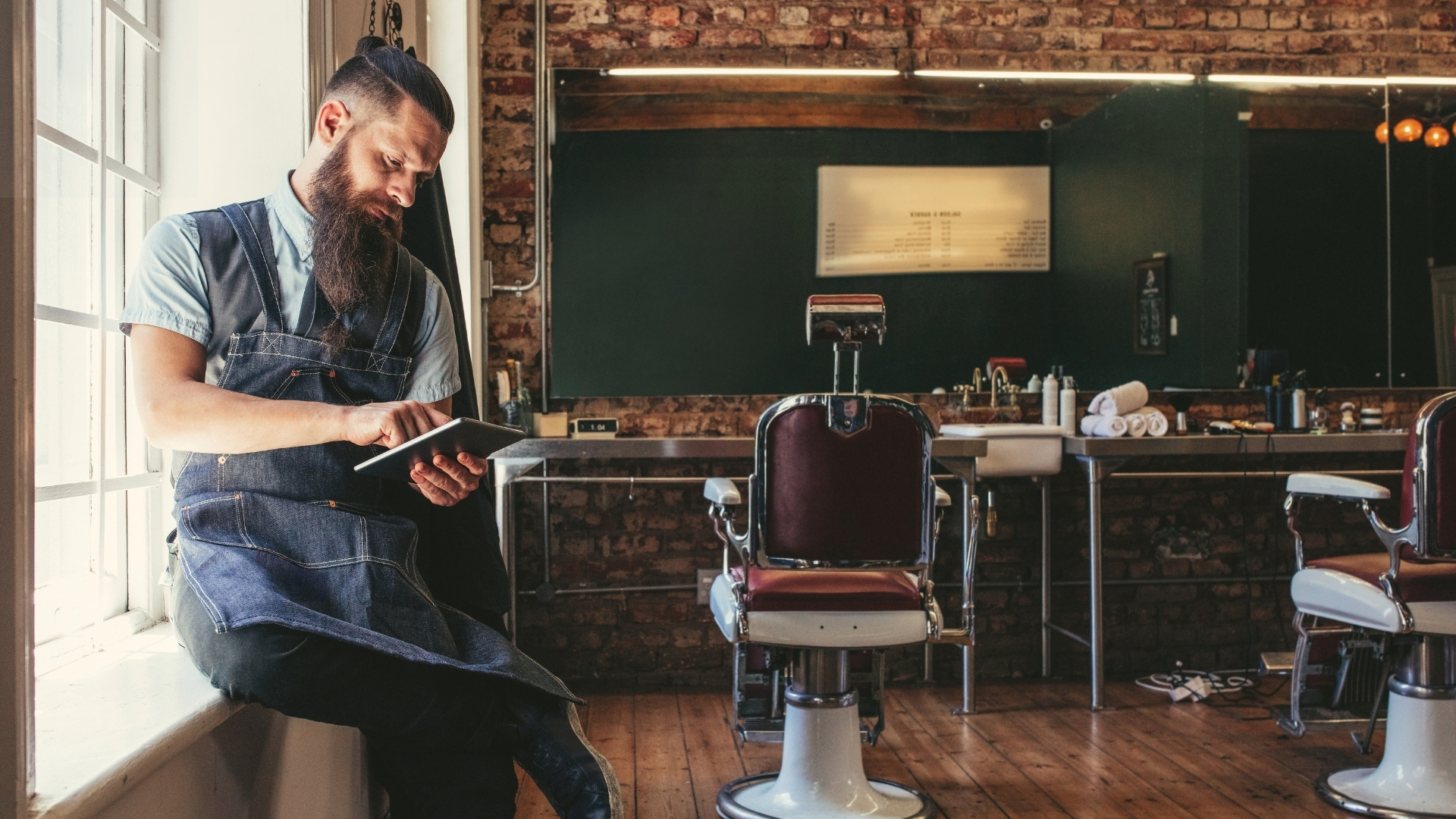 Barber relaxing by the window of his own shop, checking reviews on a tablet