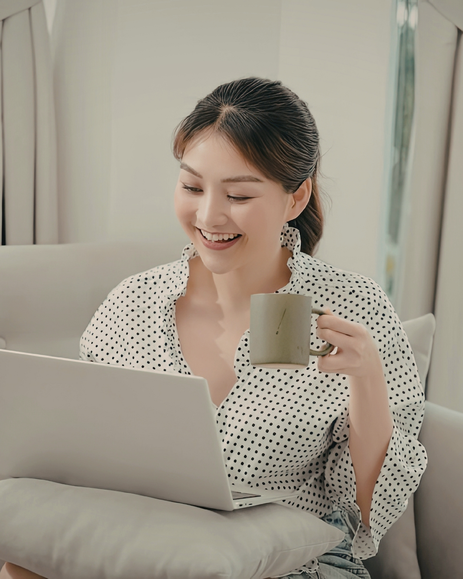 A smiling woman in a polka dot blouse sitting comfortably with a mug and laptop