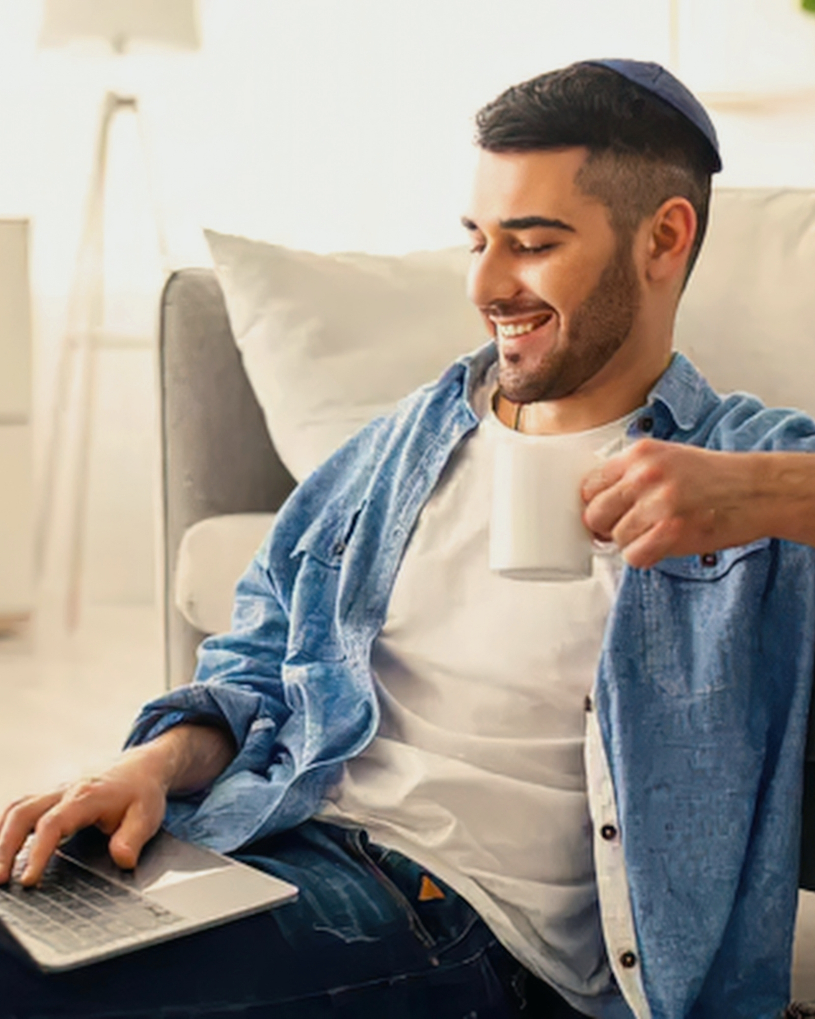 A smiling man with a mug relaxed on the floor working on his laptop