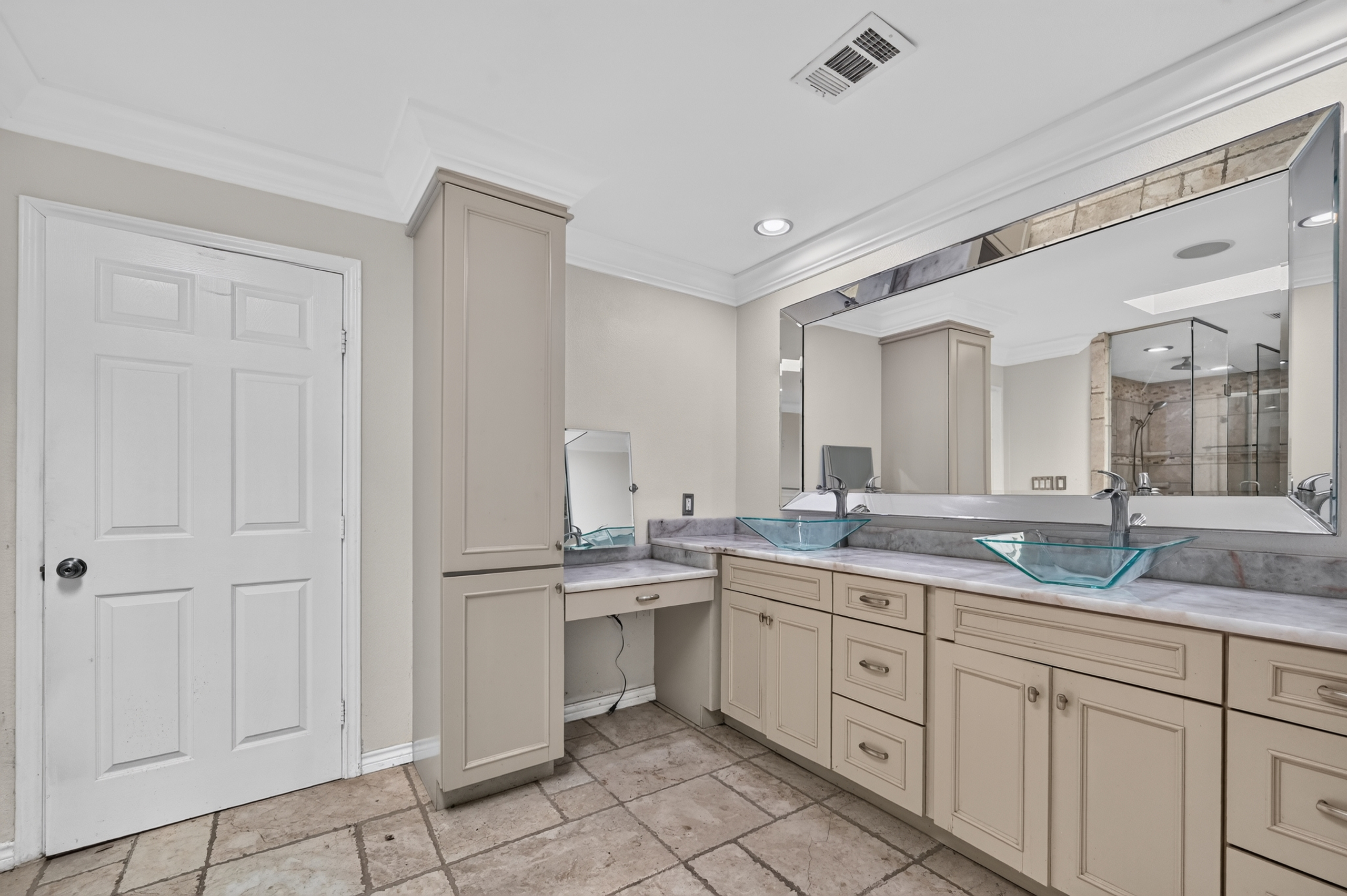 Designer luxury bathroom featuring dual glass vessel sinks, vanity makeup station, linen storage tower, and large beveled mirror