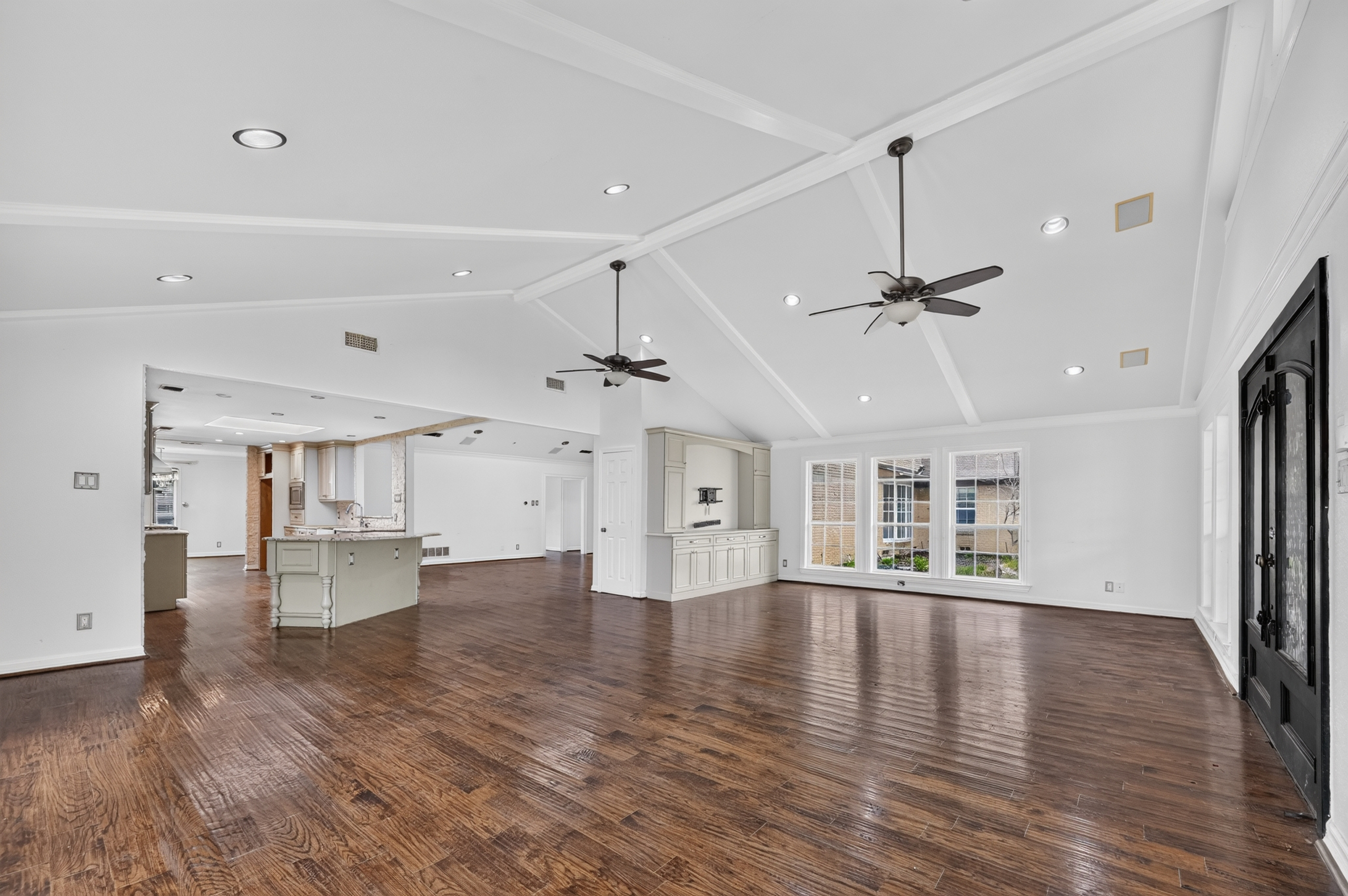 Grand open living space with soaring vaulted beamed ceiling, dual fans, hardwood floors, built-in cabinetry, and view to kitchen