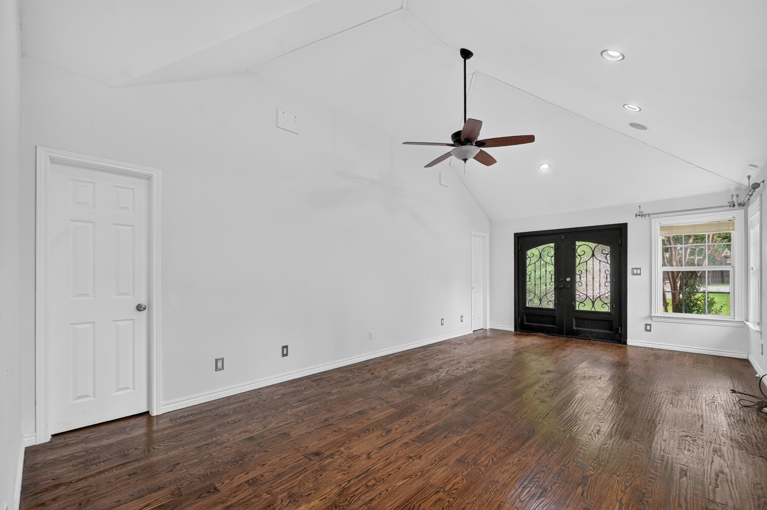Expansive vaulted great room with hardwood flooring, designer ceiling fan, and wrought-iron French doors to the grounds