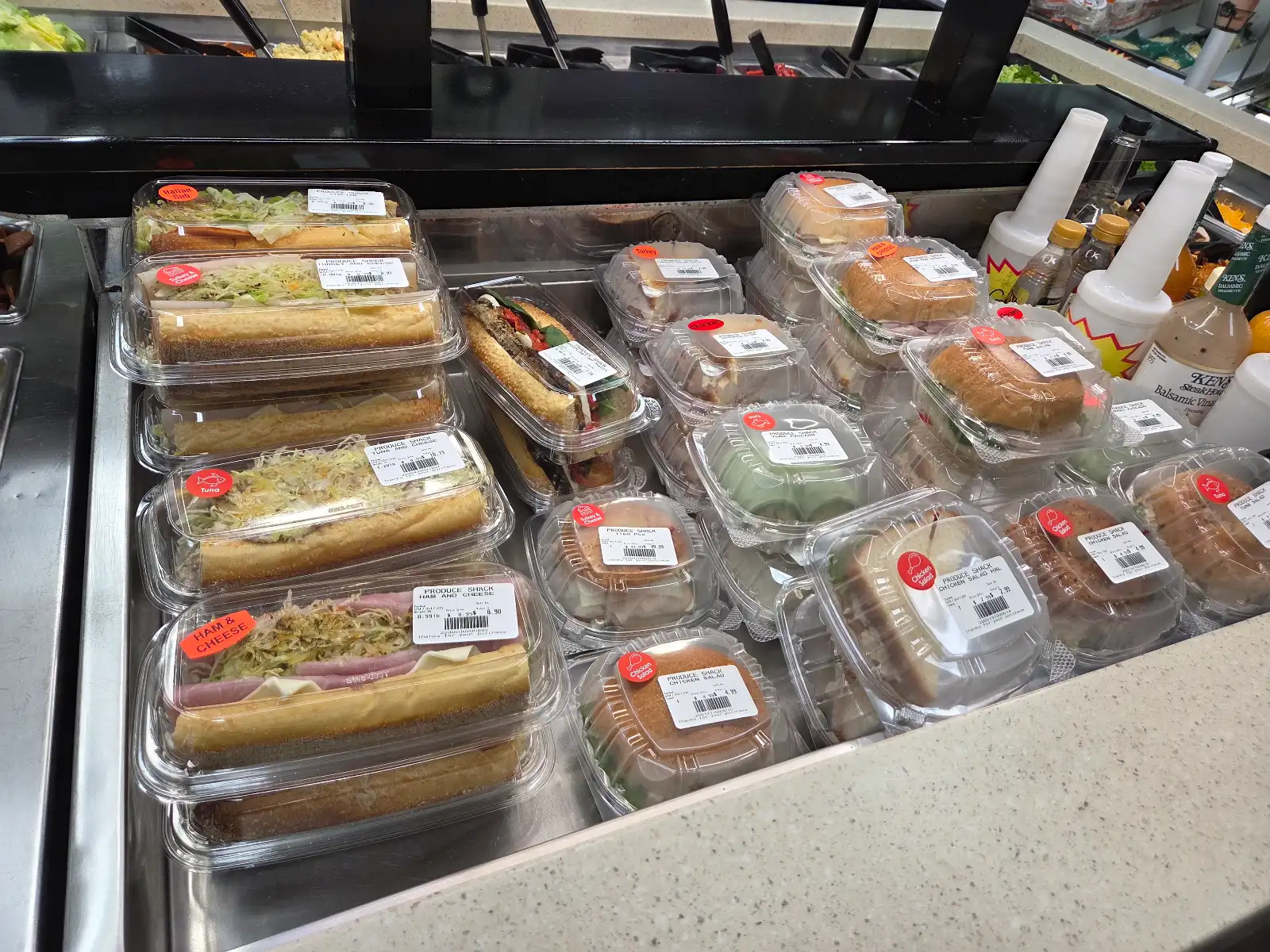 Sap's Produce Shack grab-and-go deli counter with pre-packaged Italian subs, turkey & cheese, ham & cheese, tuna, and chicken salad sandwiches