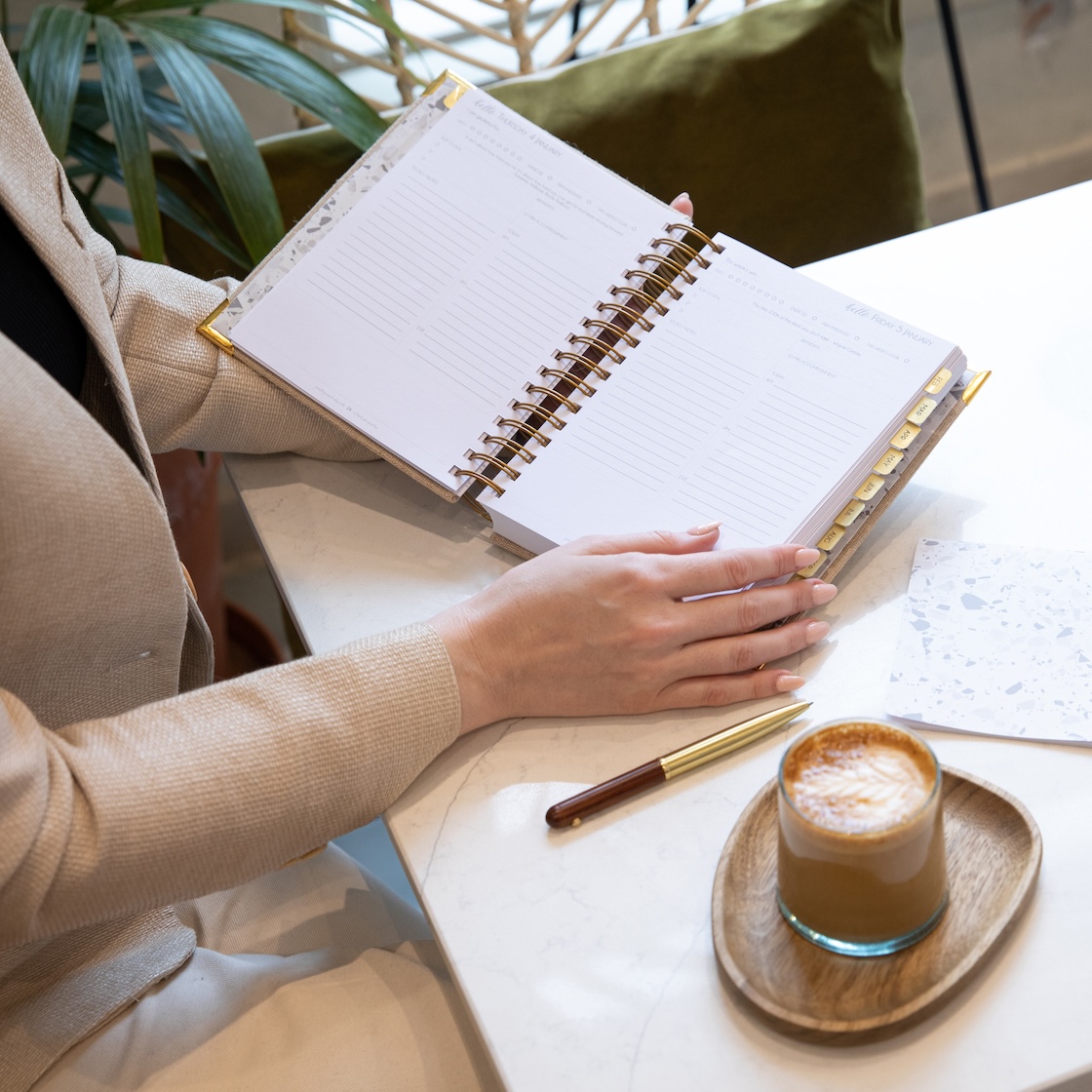 Hands holding open a spiral-bound Hello Day planner beside a coffee